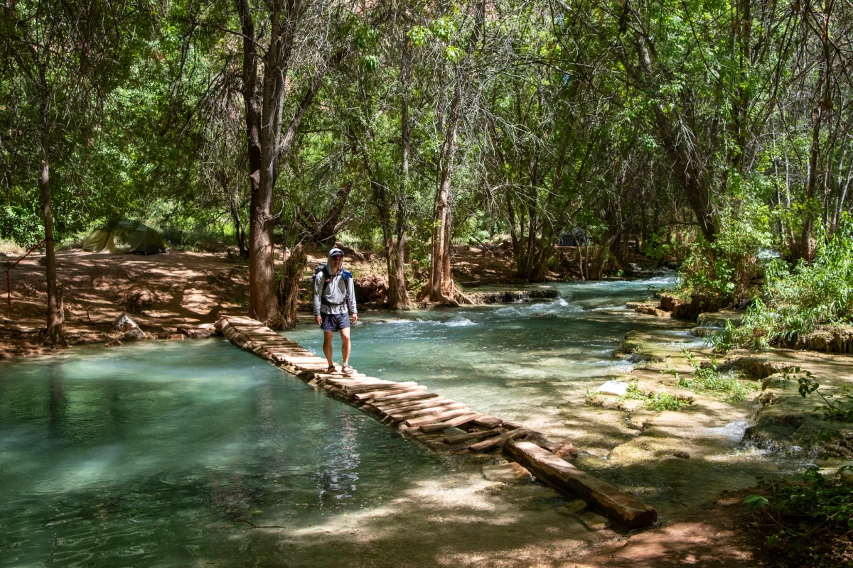 A backpacker crossing a narrow wooden plank bridge over turquoise Havasu Creek in the shade of cottonwood trees