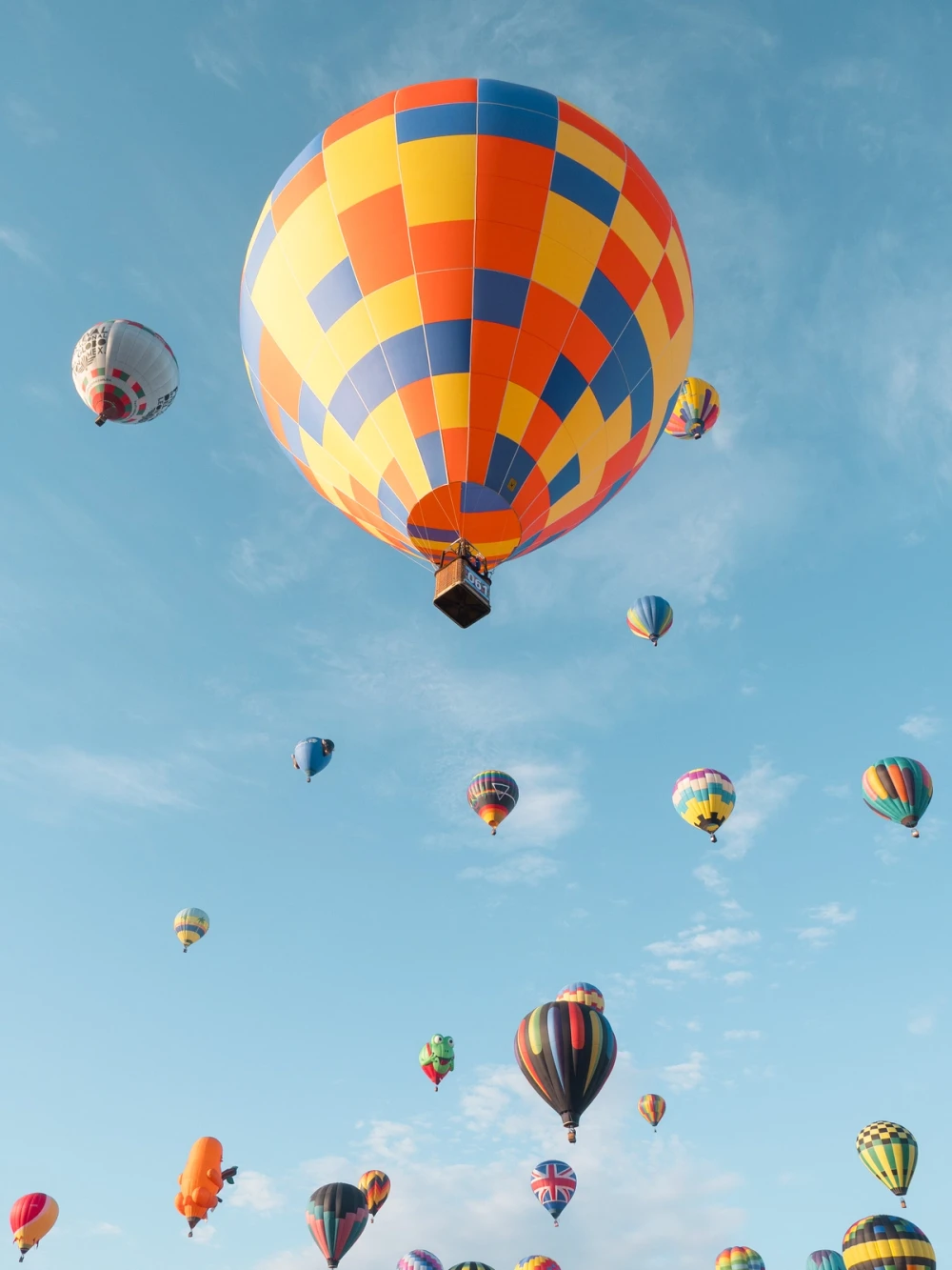A large yellow, orange, and blue checkered balloon aloft amid many smaller balloons and special shapes against a blue sky