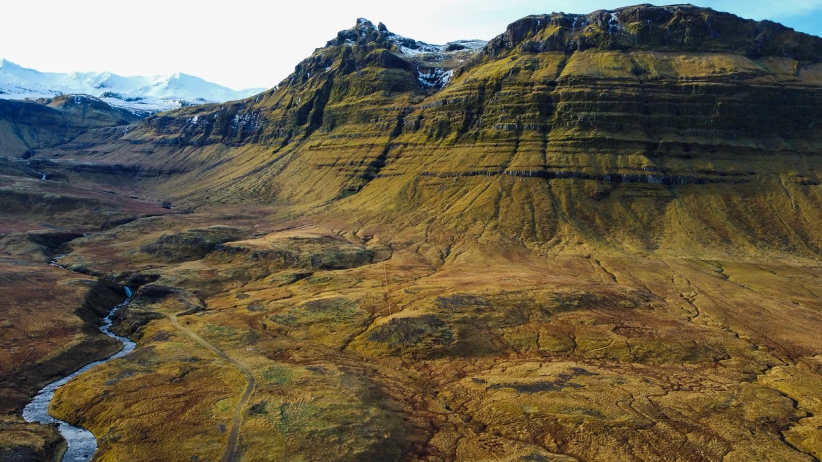 Aerial view of a layered green-and-gold Icelandic mountain cut by a winding river, snow on the ridgeline