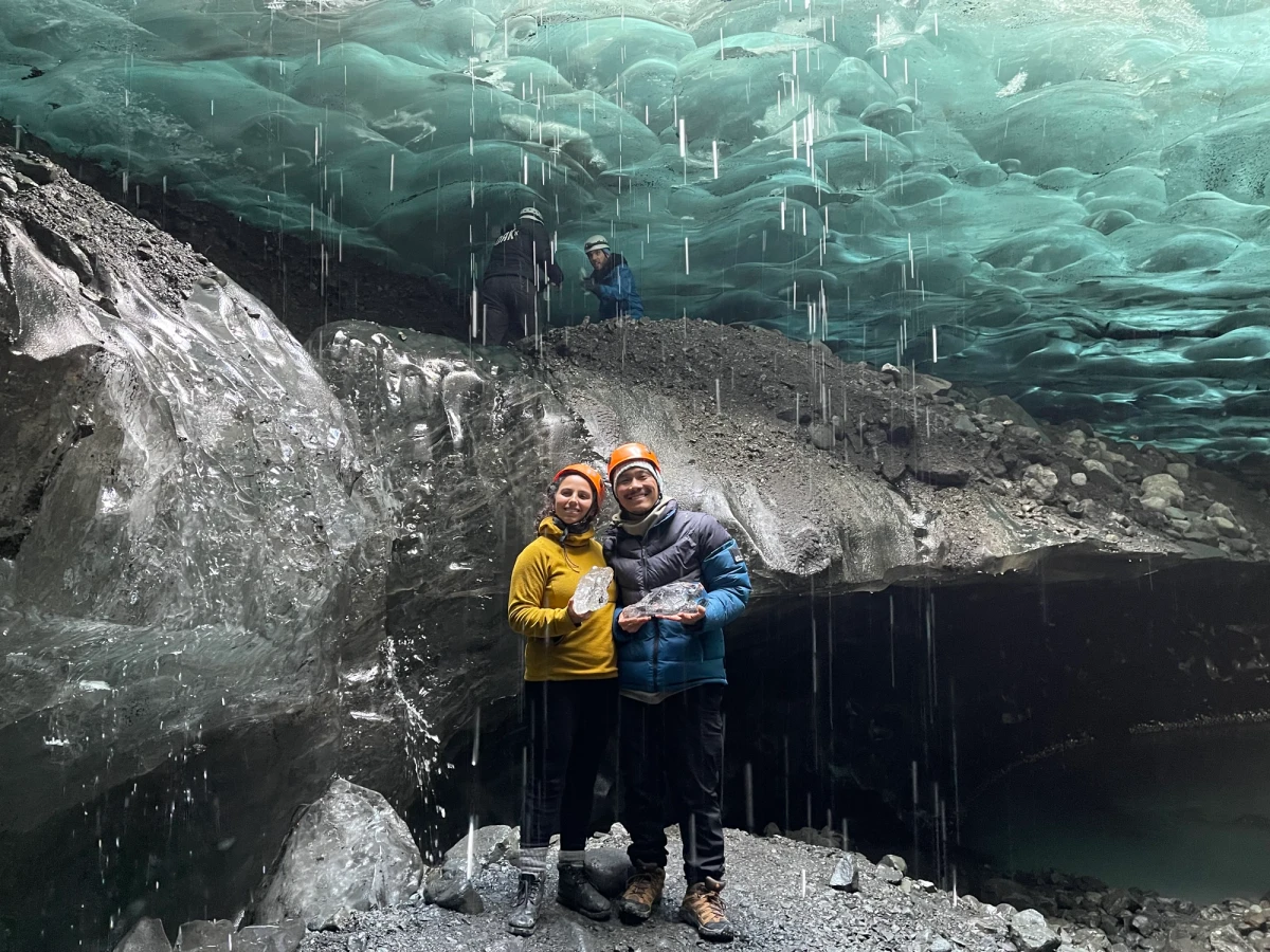 Two people standing inside a blue ice cave under a glacier, with guides and dripping meltwater