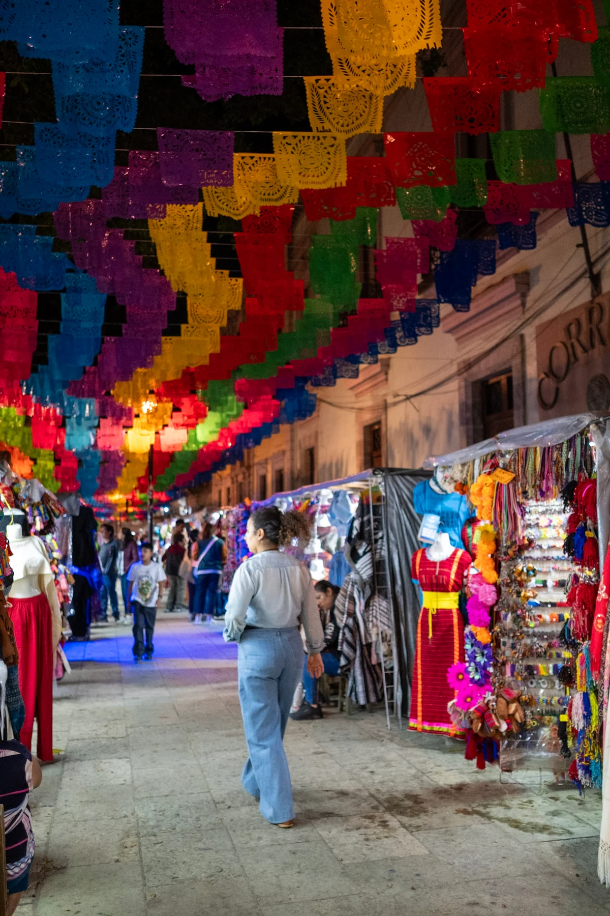 A narrow Oaxaca market street strung overhead with rows of brightly colored papel picado banners as people pass the lit stalls below