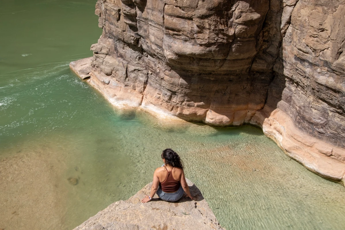 A person sitting on a rock ledge where the clear turquoise Havasu Creek meets the deeper green Colorado River beneath a tall red canyon wall