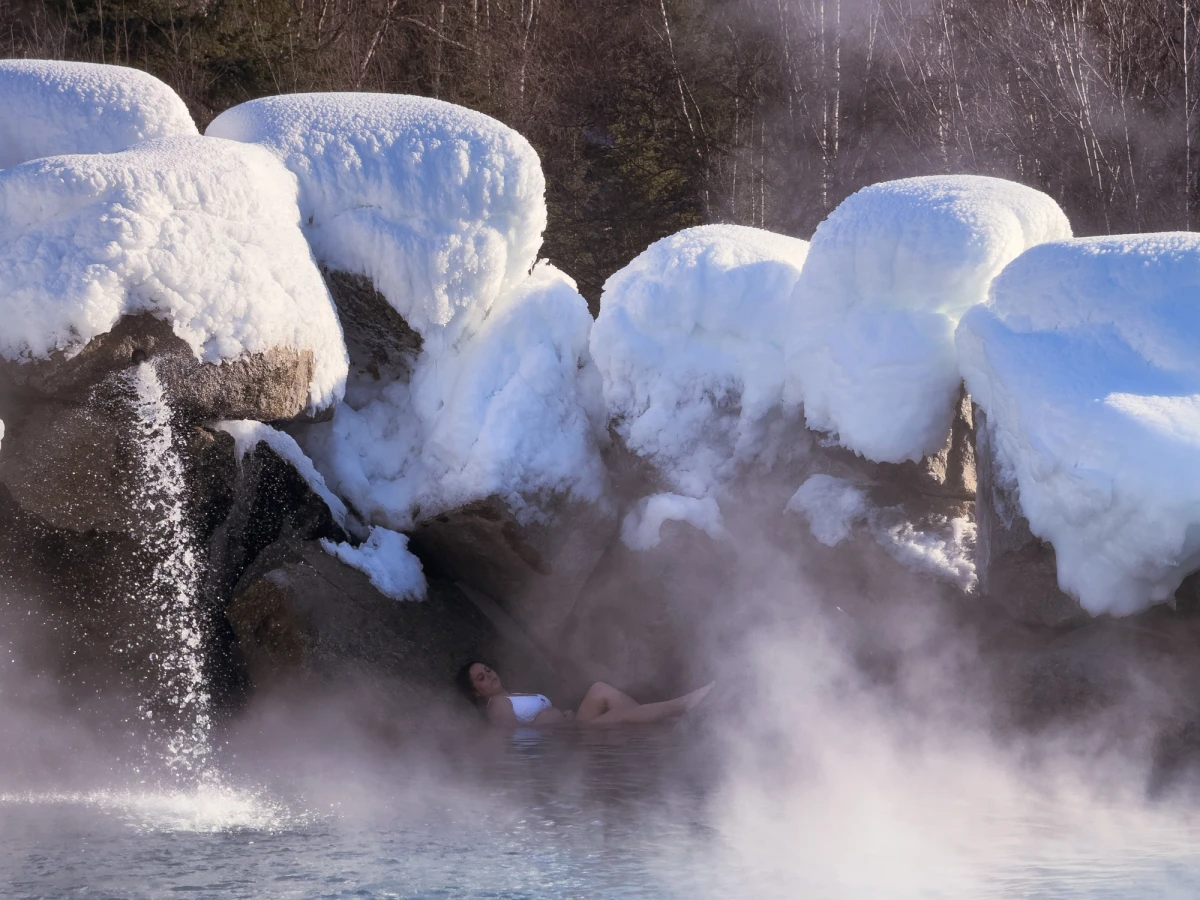 A person reclining in the steaming water of the Chena Hot Springs rock lake, ringed by large boulders capped with thick snow and rising clouds of steam