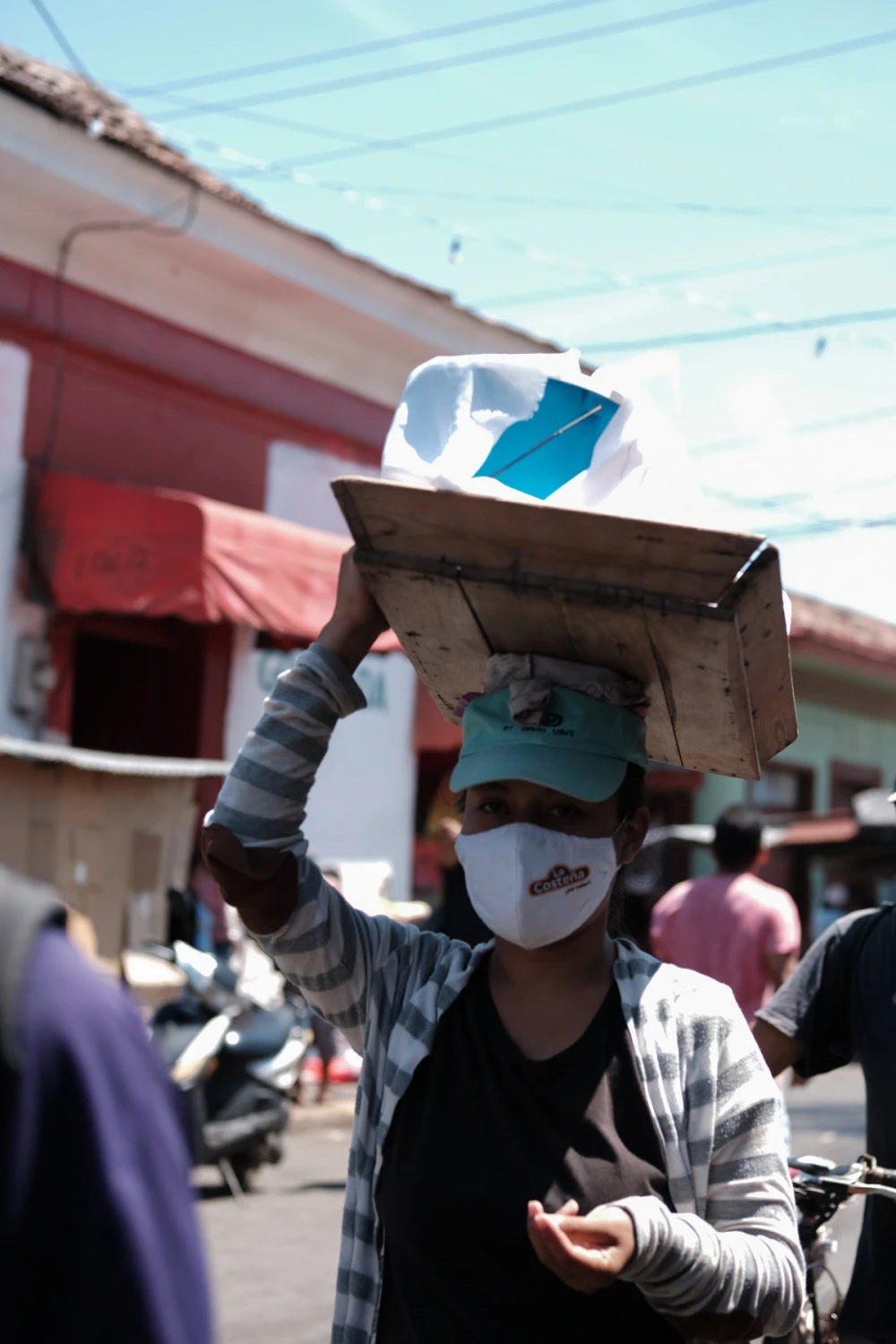 A young vendor in a face mask carrying a wooden tray balanced on their head through the market, a red colonial building behind