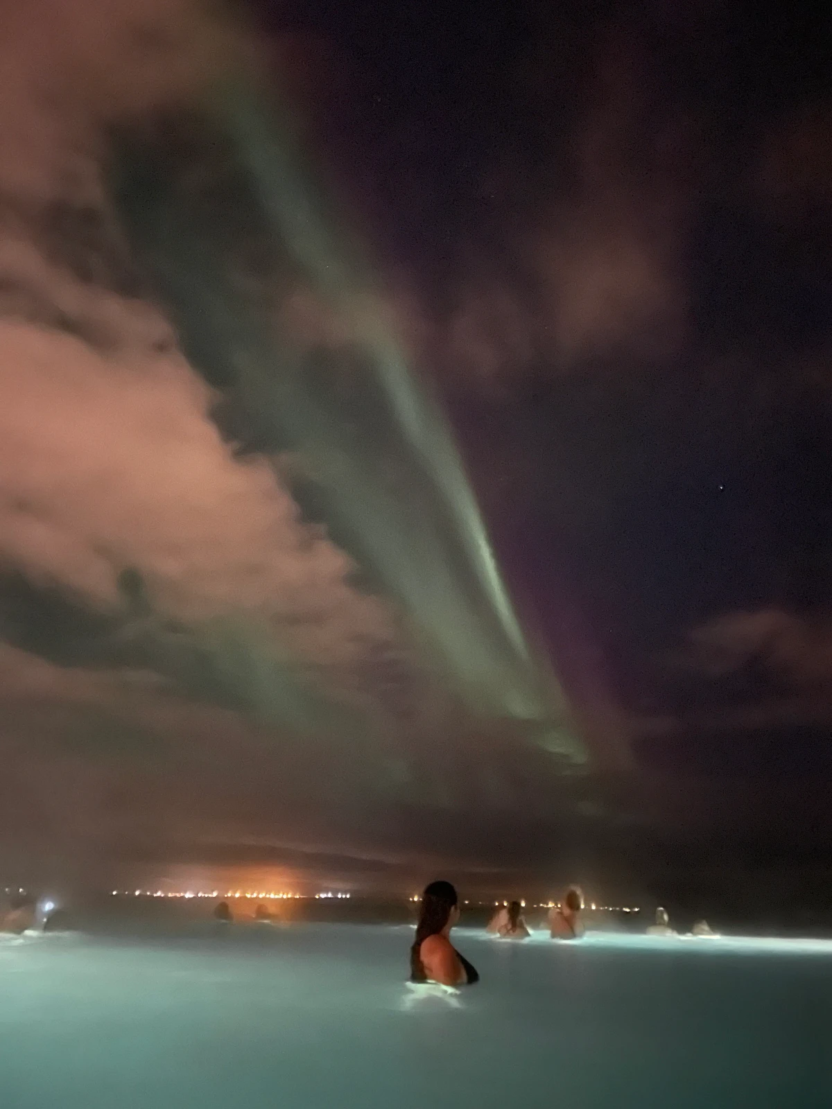 People in the Sky Lagoon geothermal pool at night with a green band of aurora in the clouds and town lights on the horizon