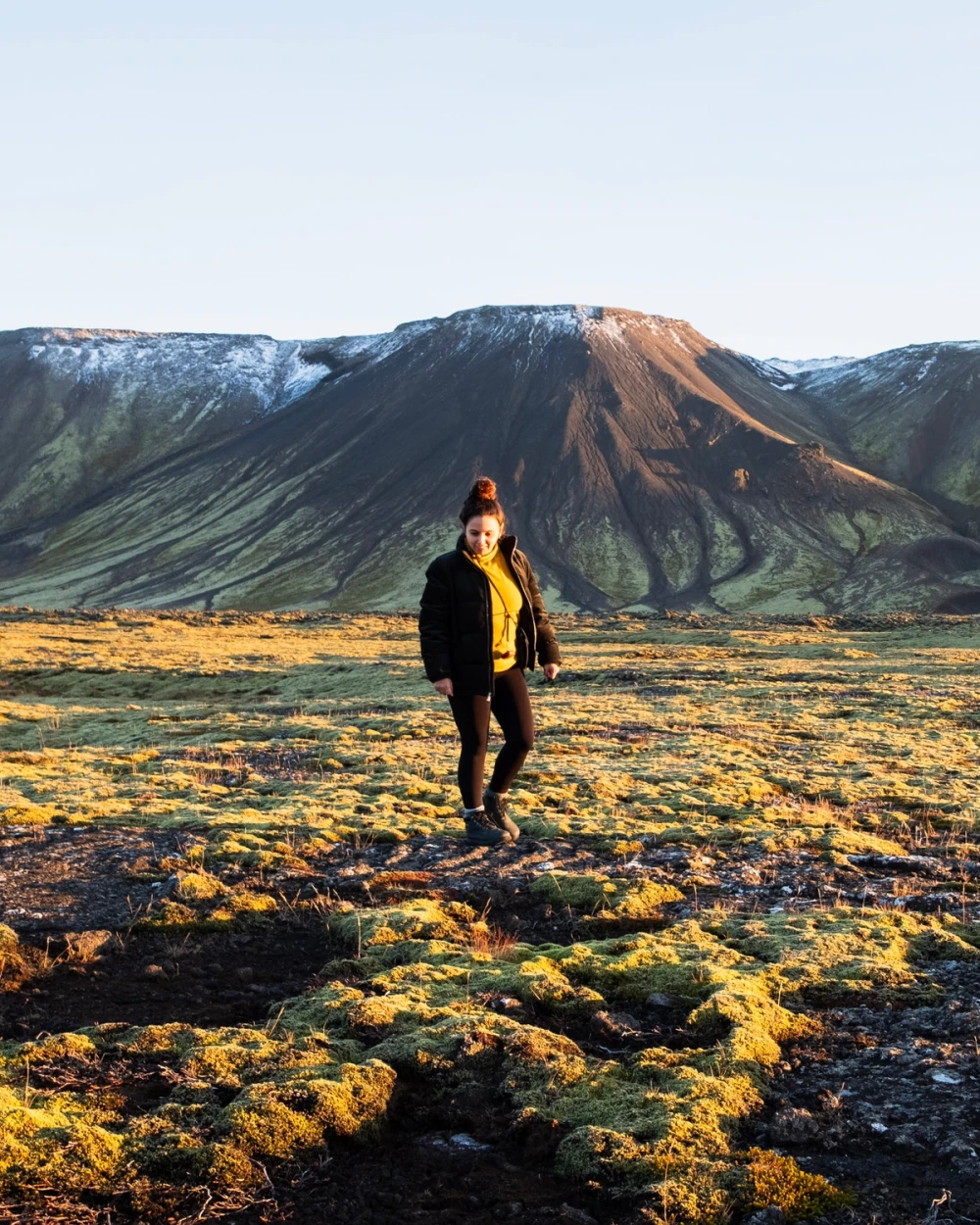 A person standing in a mossy field in golden light below a snow-dusted volcanic cone