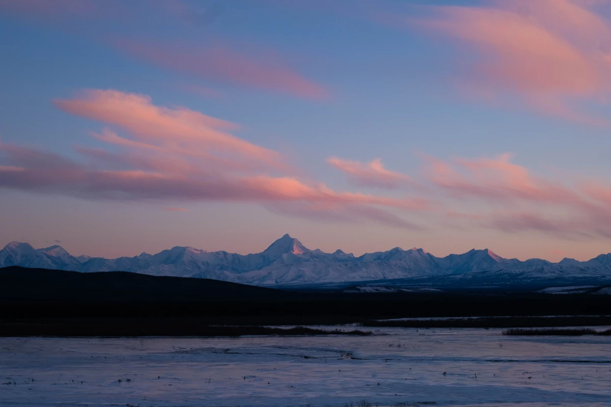 The snow-covered Alaska Range catching pink dusk light beneath streaked clouds, frozen flats in the foreground