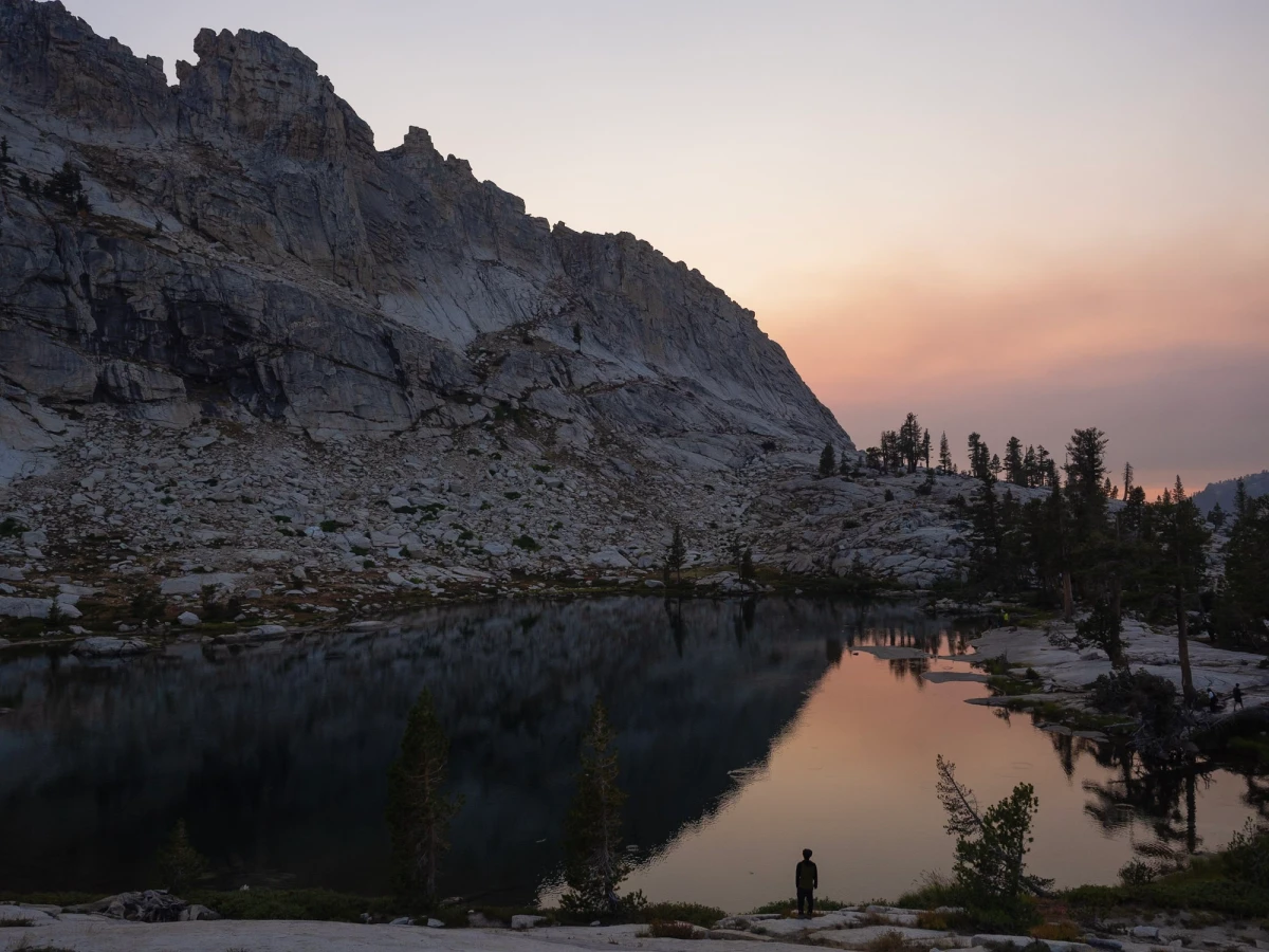 A still alpine lake at dusk reflecting a towering granite cliff, with a small lone figure standing on the far shore