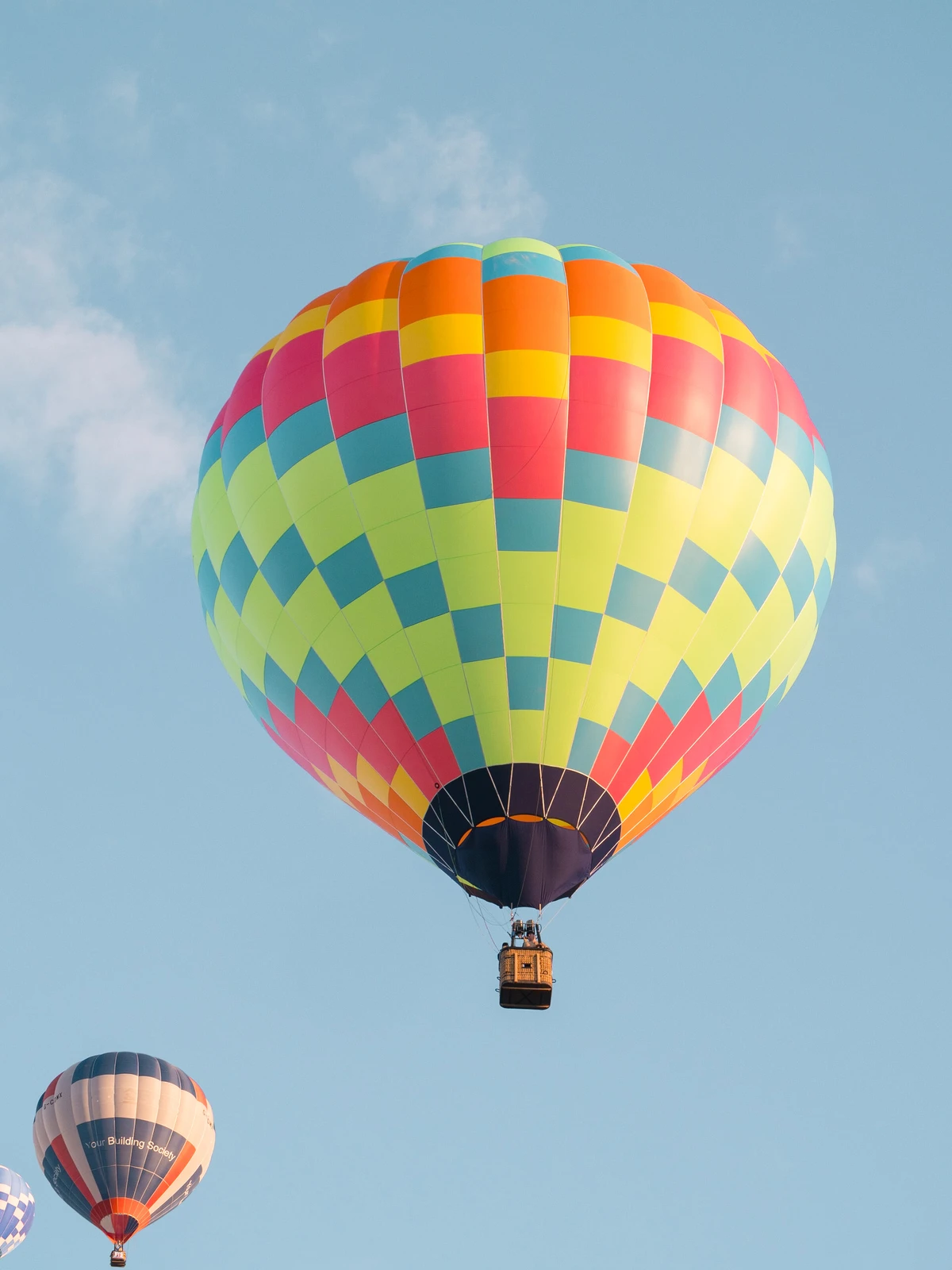 A single multicolored checkered hot-air balloon rising into a clear blue sky with two more balloons below it