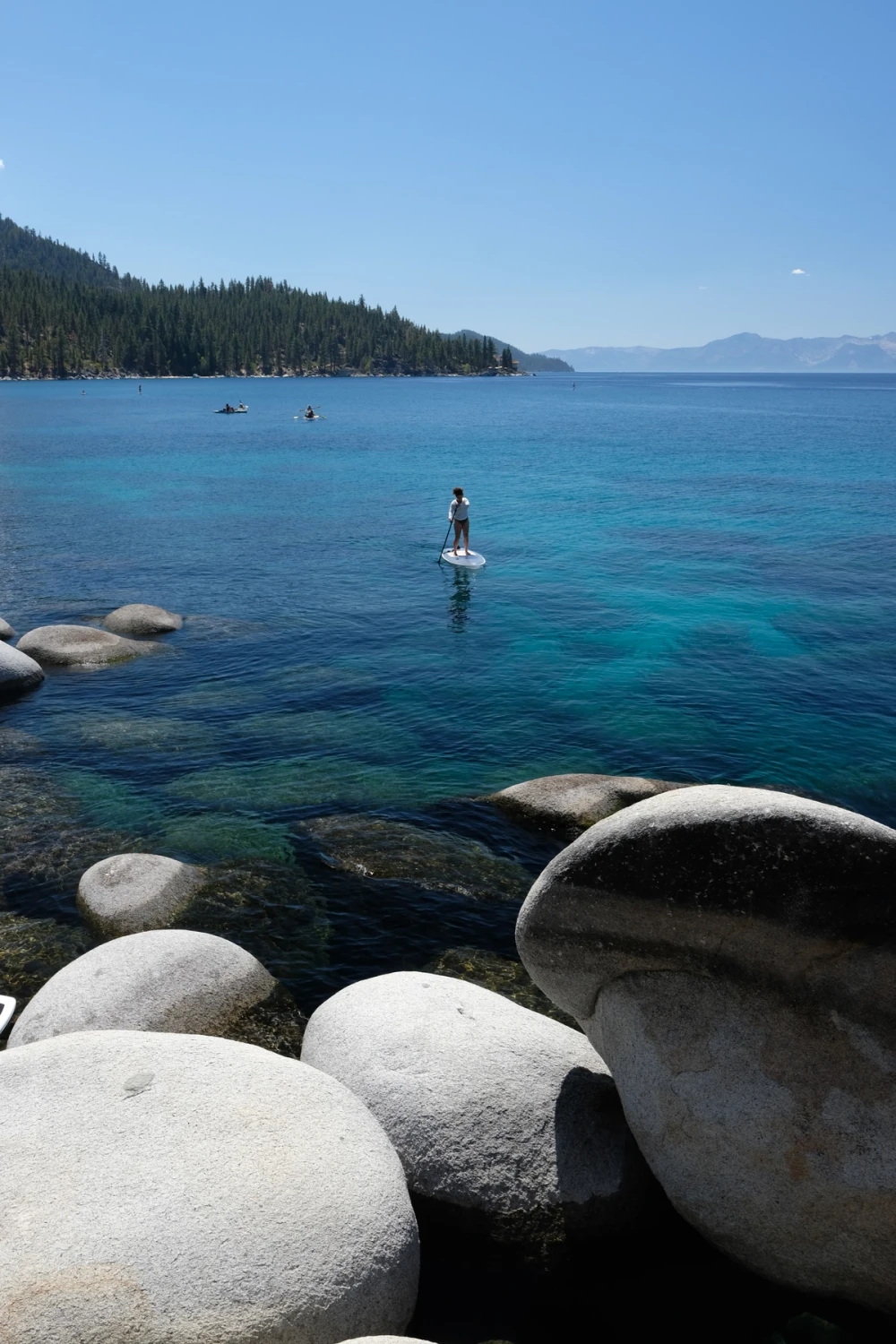 A person standing on a paddleboard on glassy clear water among granite boulders, mountains across the lake