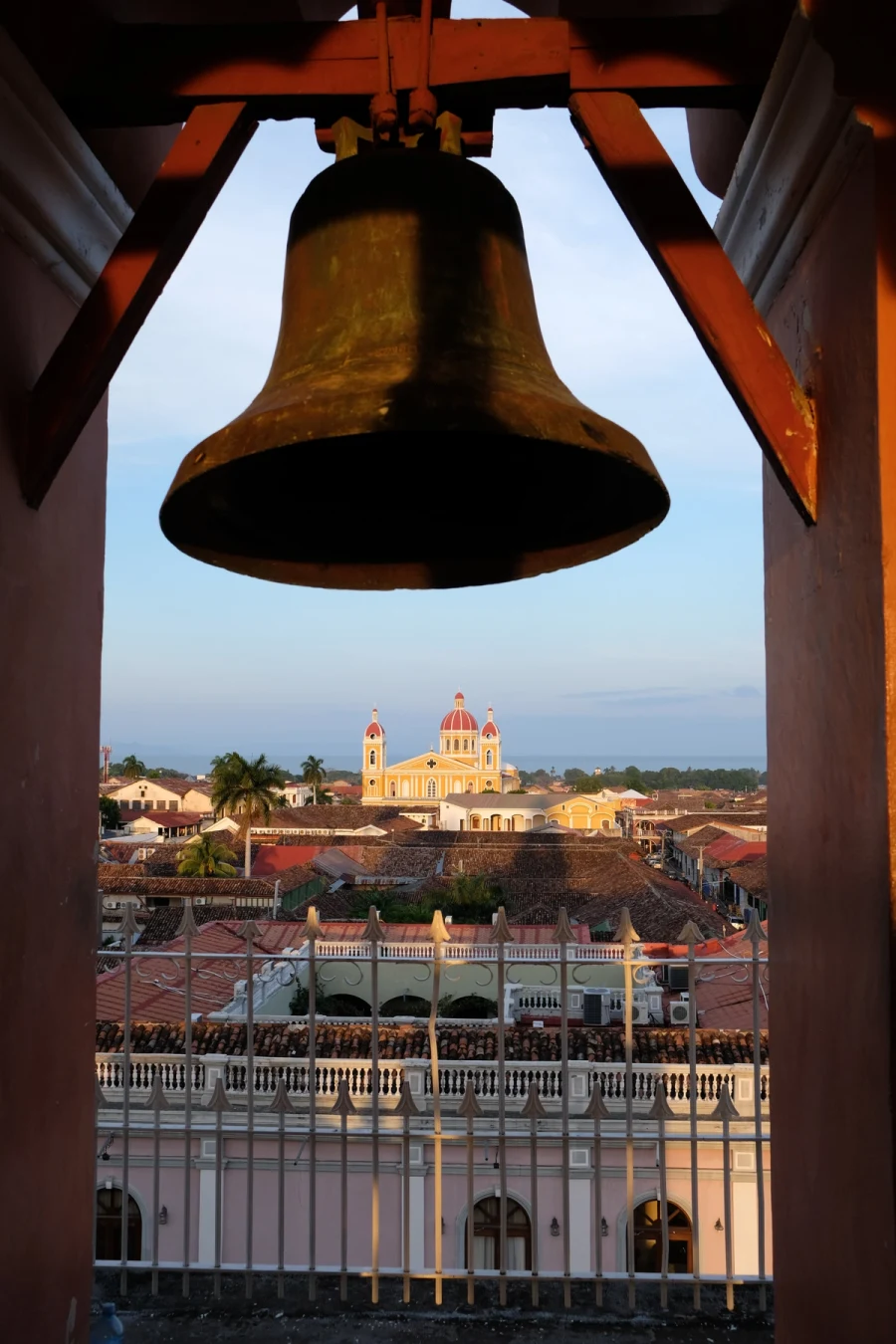 The view from a church bell tower over the tiled rooftops of Granada to the yellow Cathedral of Granada, with Lake Nicaragua and a hazy volcano beyond at golden hour