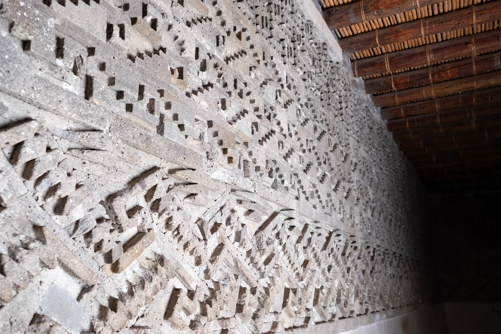A long wall of intricate geometric stone fretwork at Mitla, raking light catching the relief, a reed ceiling above