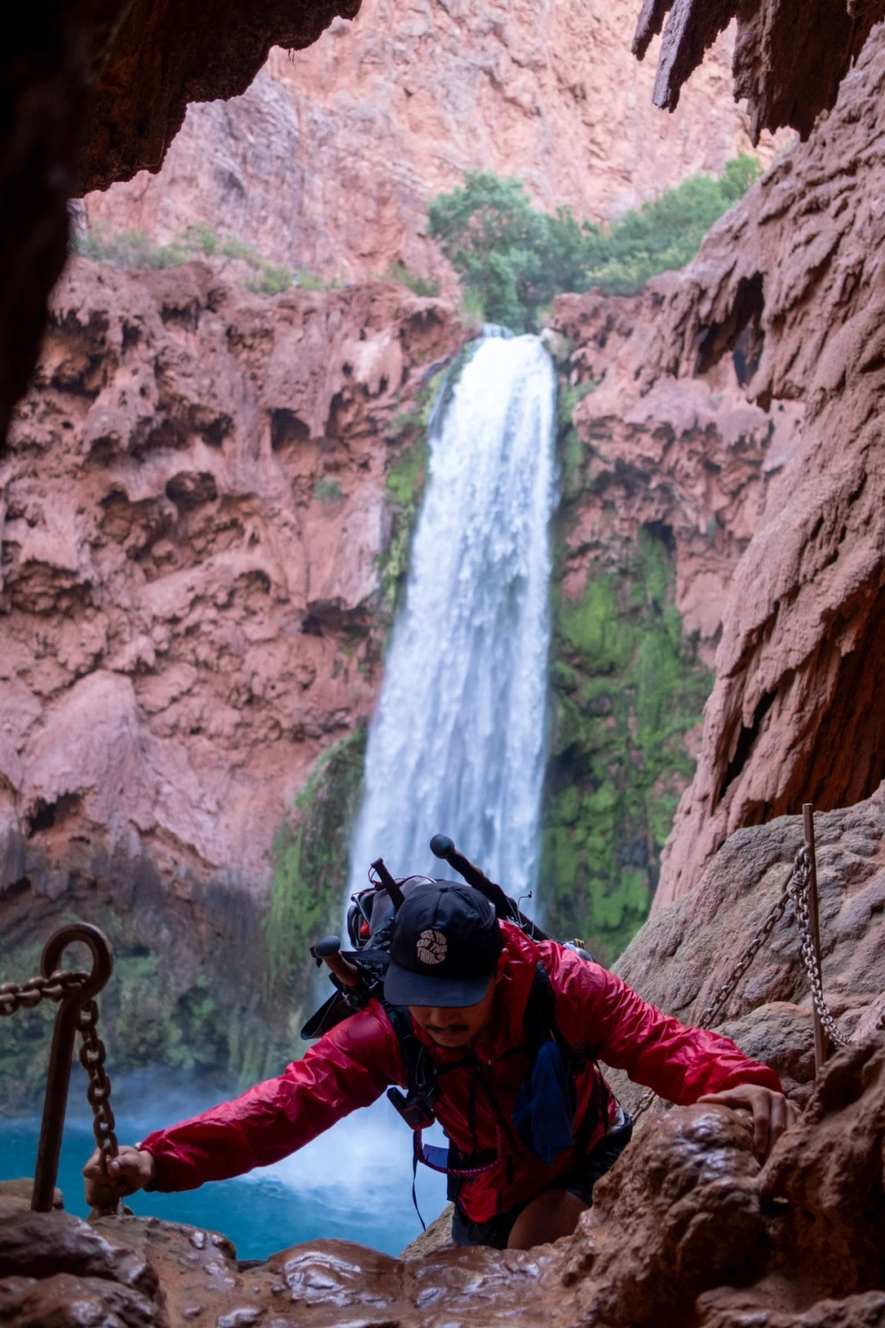 A hiker in a red jacket gripping a chain on the steep, wet descent beside Mooney Falls, the waterfall plunging into a blue pool behind