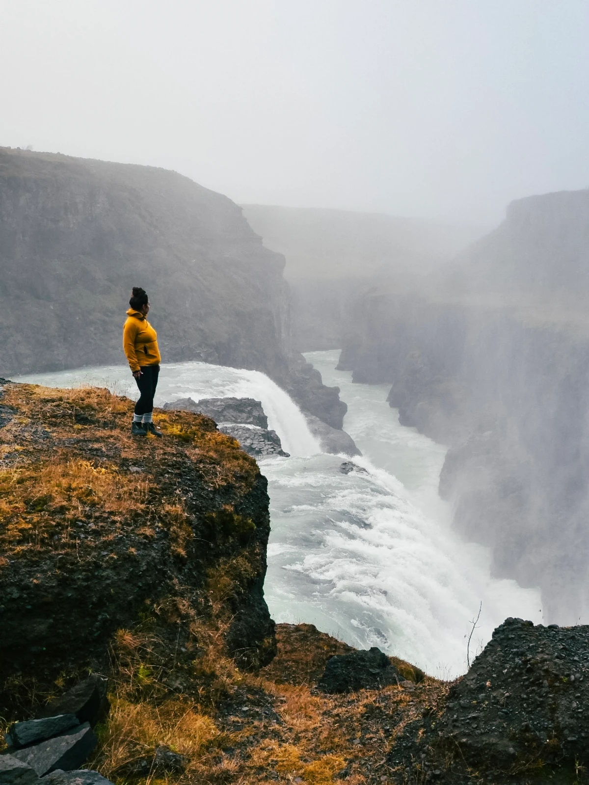 A person in a yellow jacket standing on a cliff edge above the powerful two-tiered Gullfoss waterfall in heavy mist