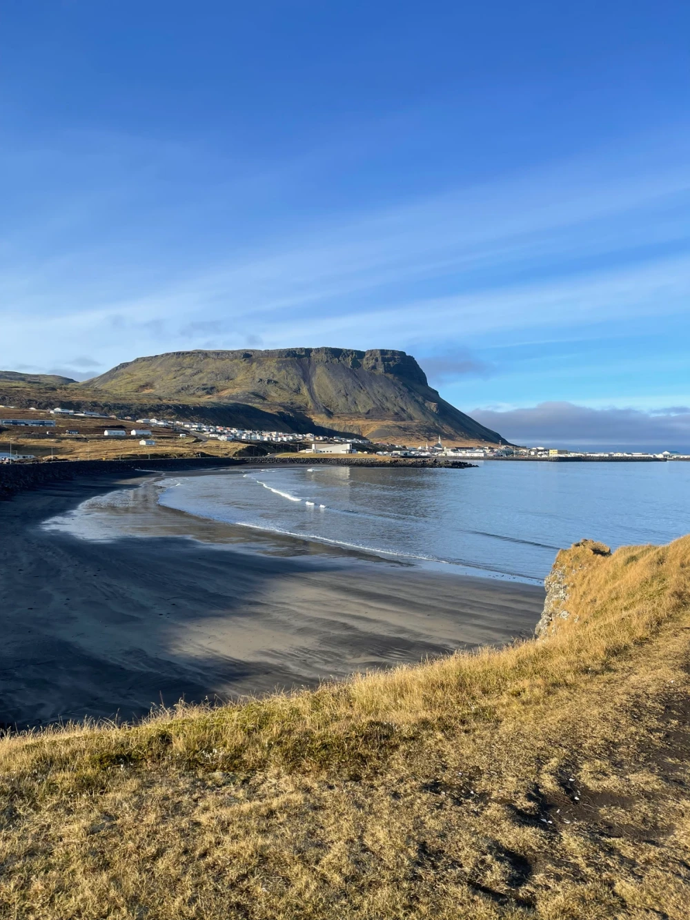 A black-sand bay curving below a flat-topped green headland and a small coastal town