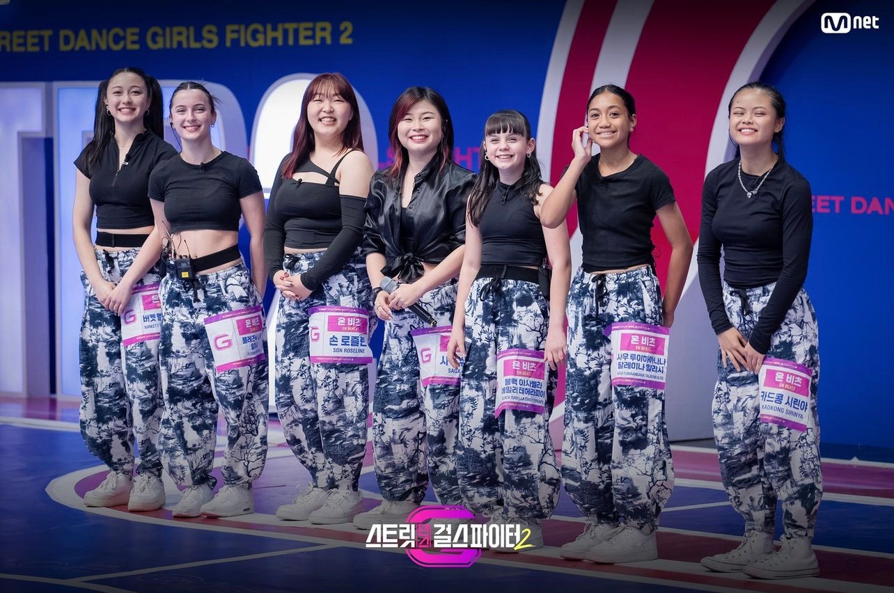 Seven dancers in matching camo pants and black shirts smiling on TV