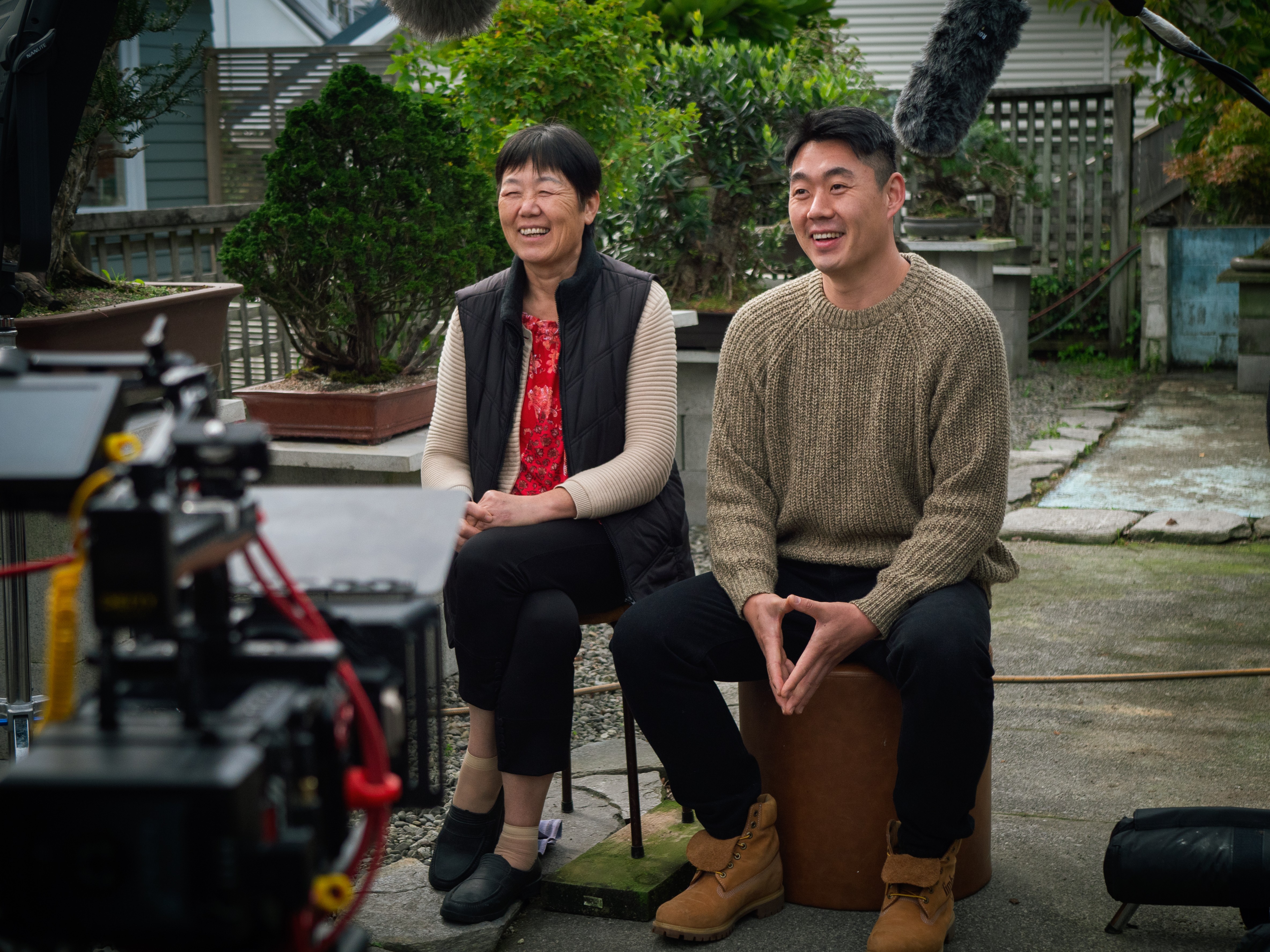 A man and his mother sit side by side in a garden, while a camera films them