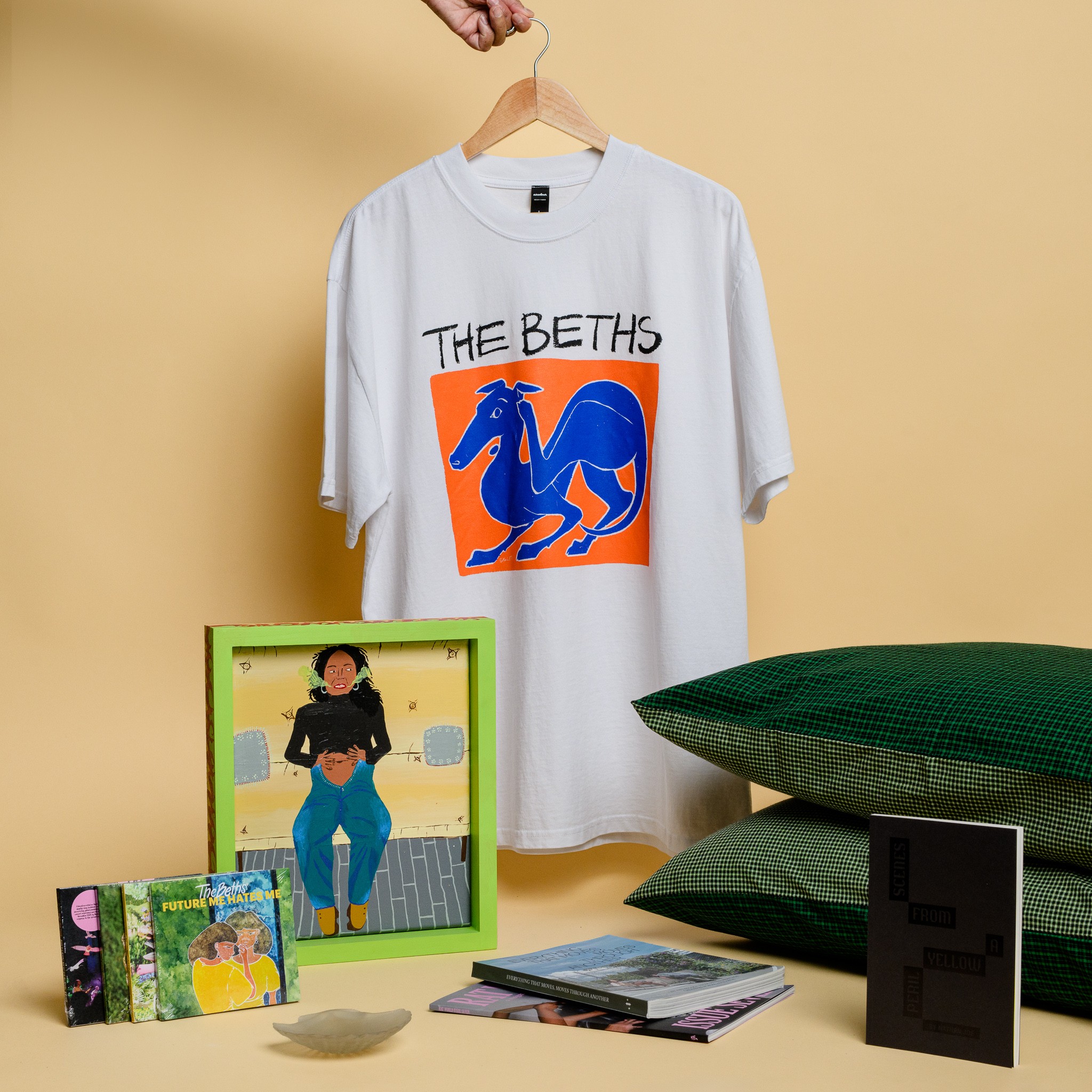 A white t-shirt with and orange and blue print of a dog, a painting of a woman clutching her gut, a set of CDs, a glass scallop, two green pillowcases and a pile of three books