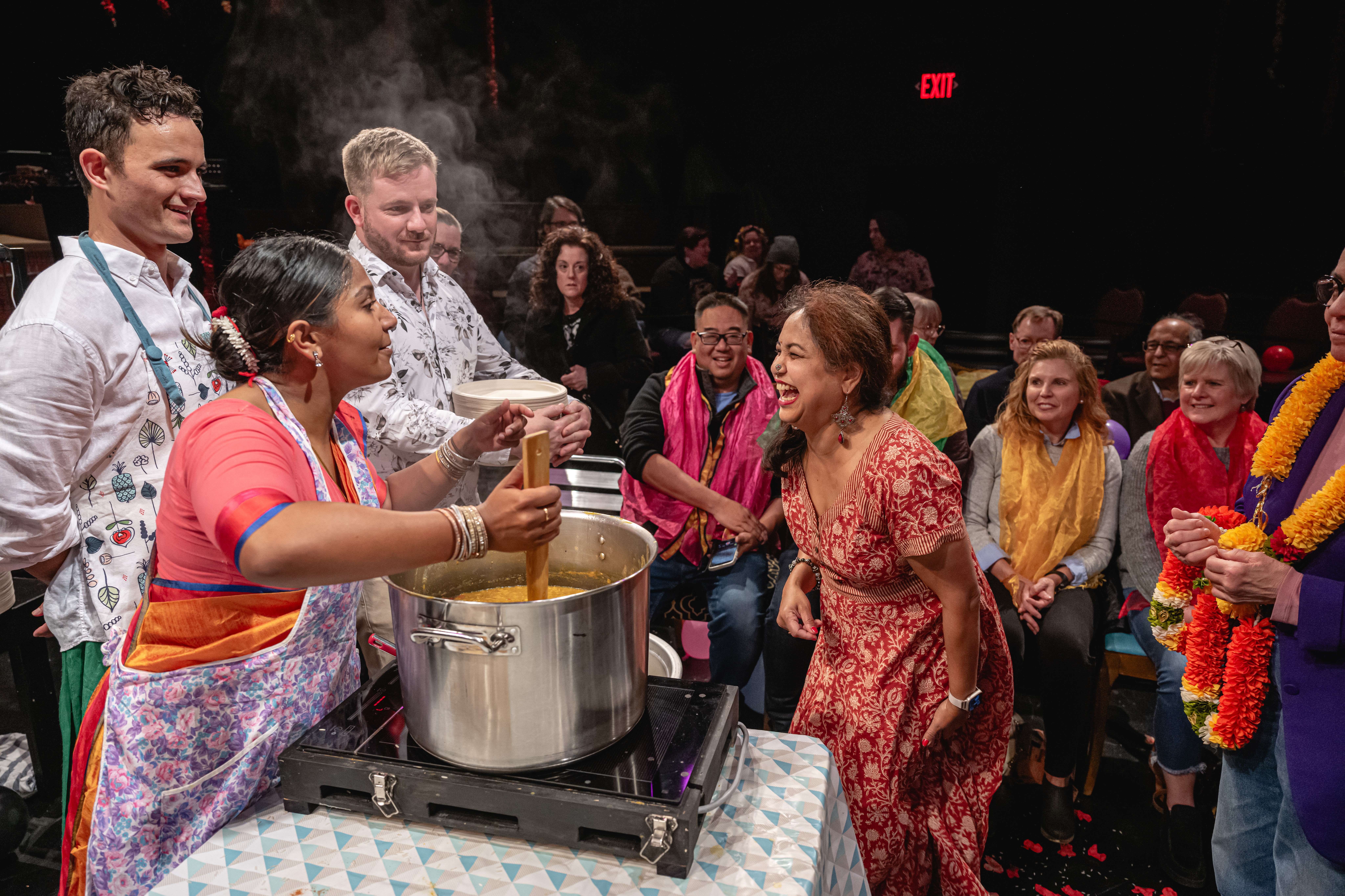 A group of people in aprons are gathered around a large silver pot while an audience member approaches the table