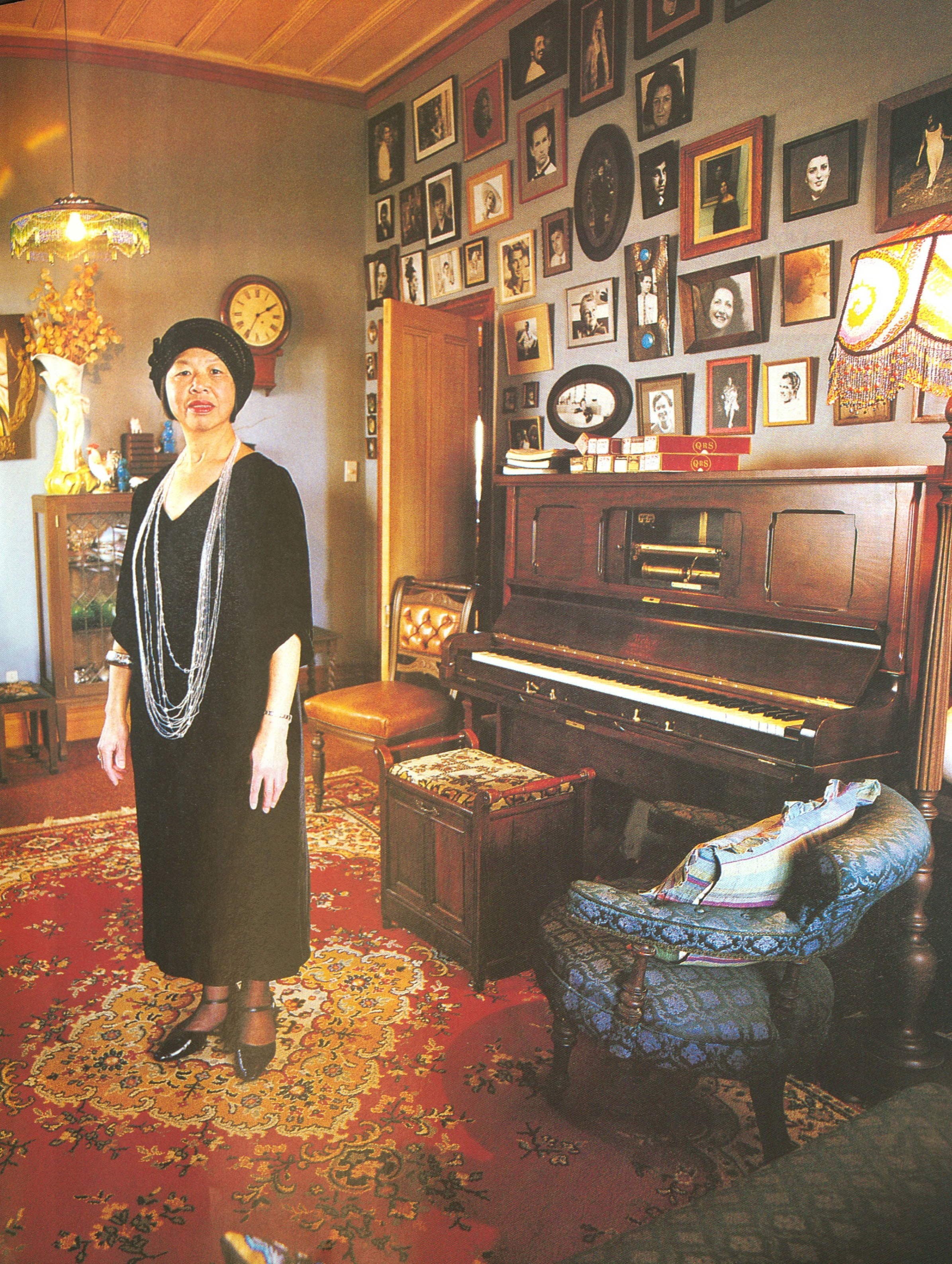 A woman in vintage clothes standing in a room with a piano, upholstered chair, and framed photographs on the wall