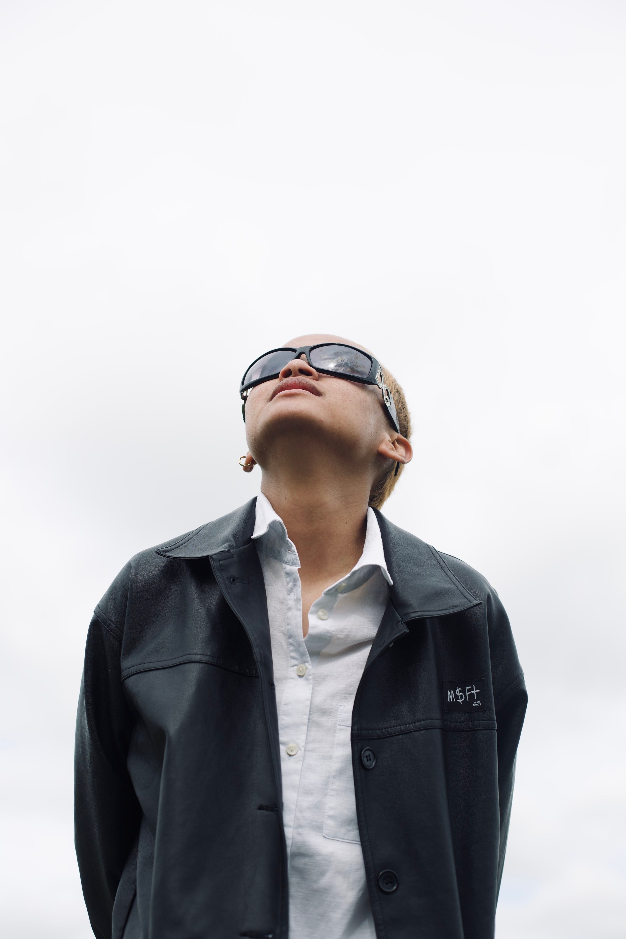 Filipino person looking up at the sky wearing huge mirrored sunnies, a button-down shirt and dark jacket