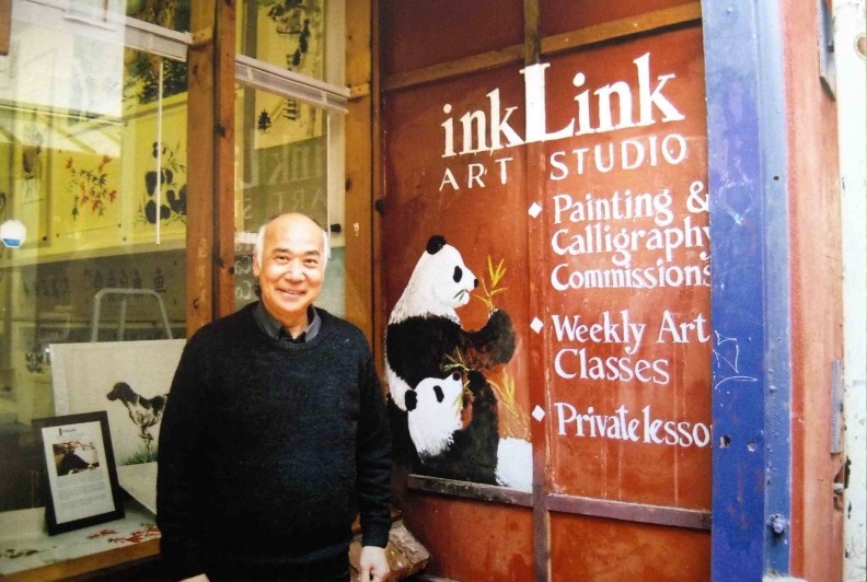 A smiling man stands outside an art studio, in front of a painted wall advertising classes and paintings with a panda mural