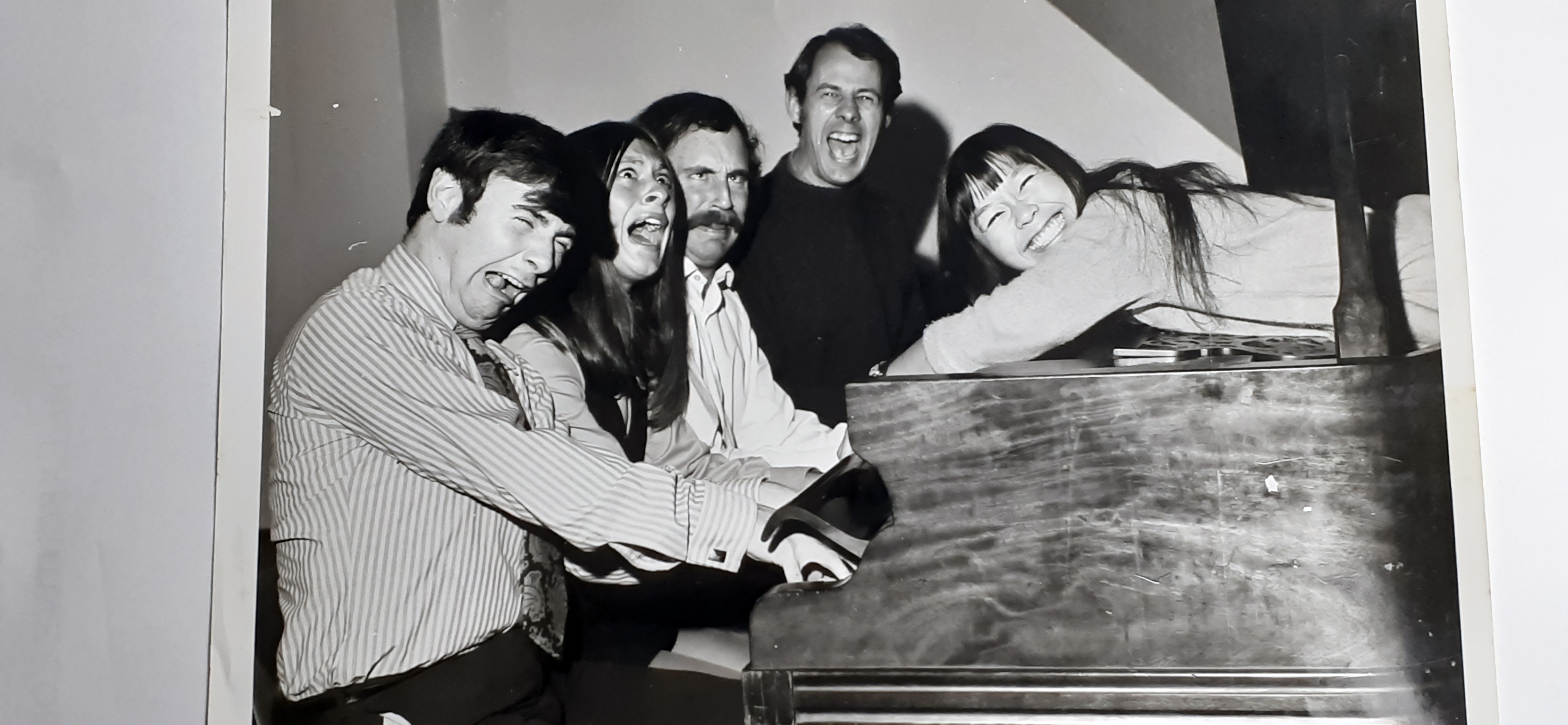 Three men and one woman sit at a piano, singing a song. A second woman, smiling, lies on top of the piano.