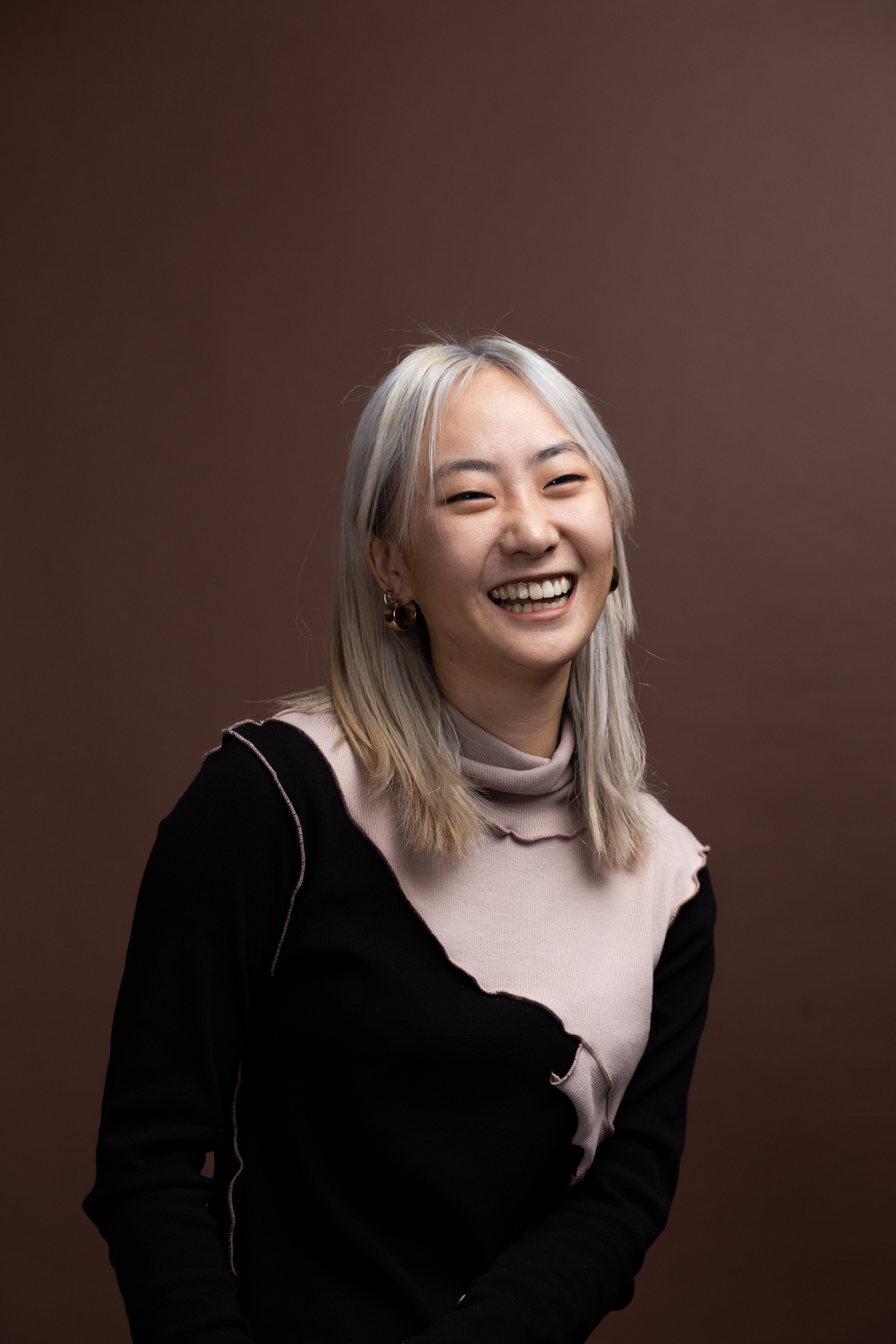Nahyeon has mid-length silver hair and is laughing for the camera in front of a dark background.