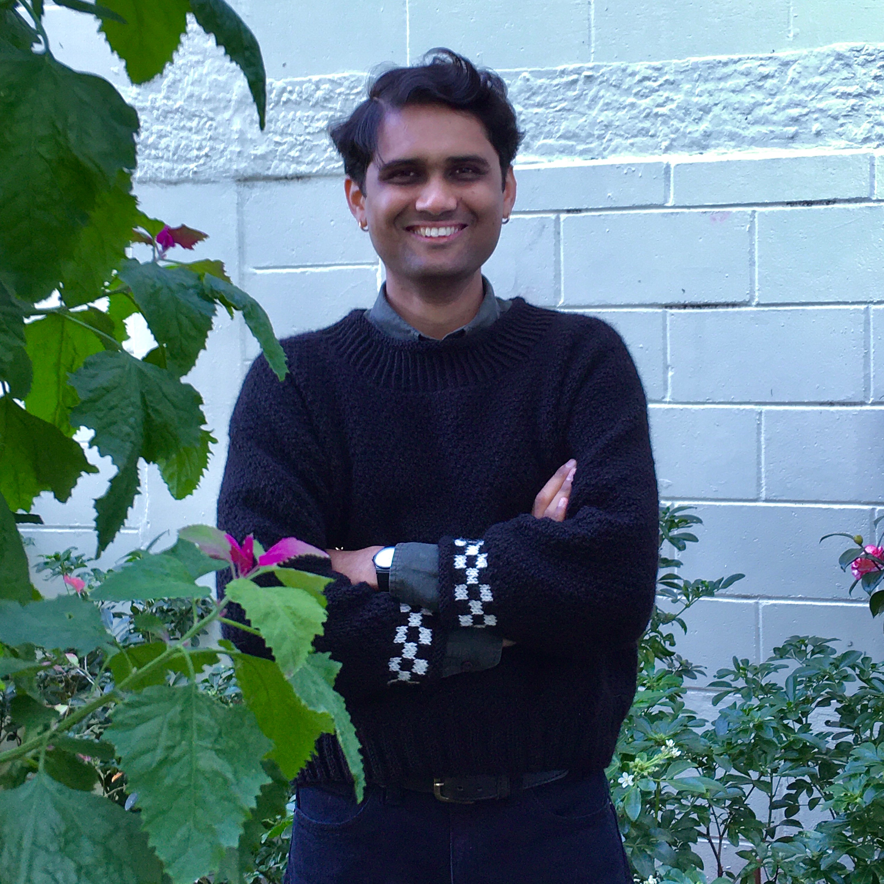 Photo of a man in front of a white brick background amongst flowering greenery. He is smiling, wearing jeans and a black jumper with black and white checkered cuffs.