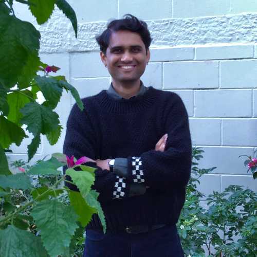 Photo of a man in front of a white brick background amongst flowering greenery. He is smiling, wearing jeans and a black jumper with black and white checkered cuffs.