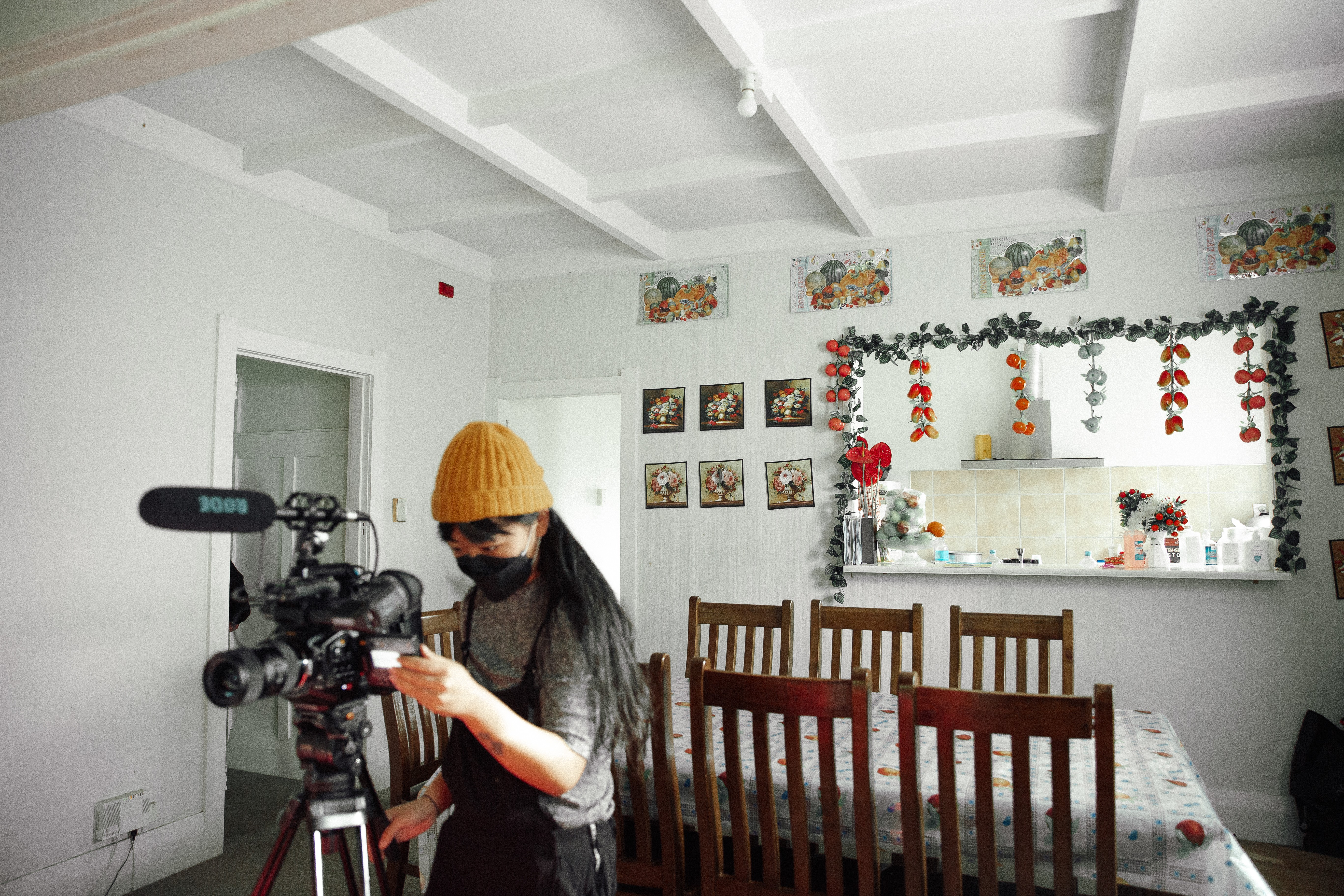 A woman in a mask and a yellow beanie standing behind a camera in a dining room