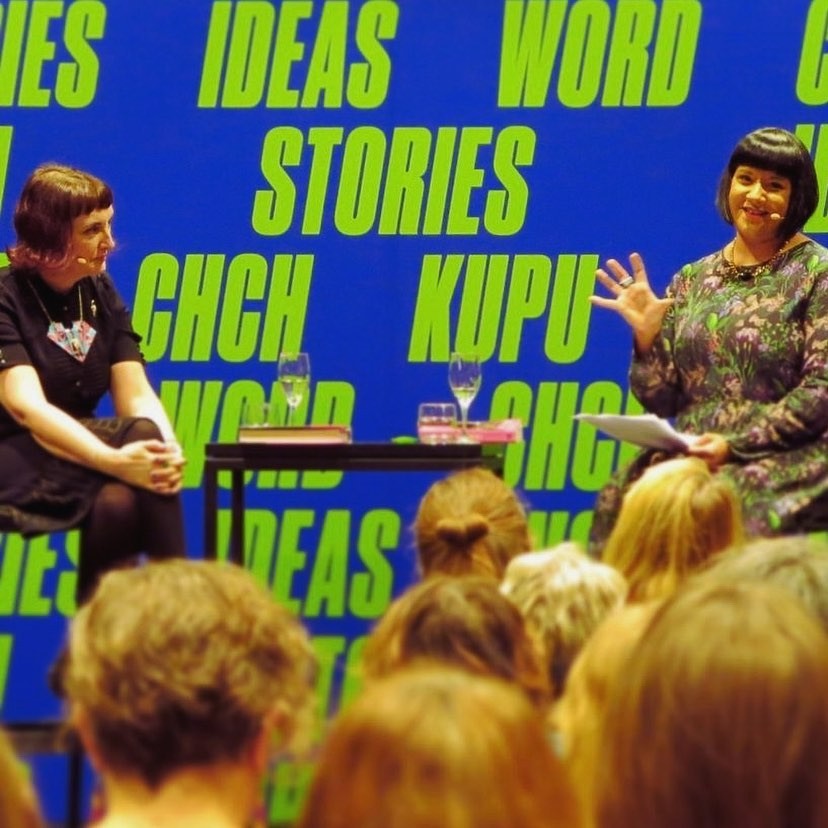 Two people sitting on a stage in front of a crowd with a blue and green background
