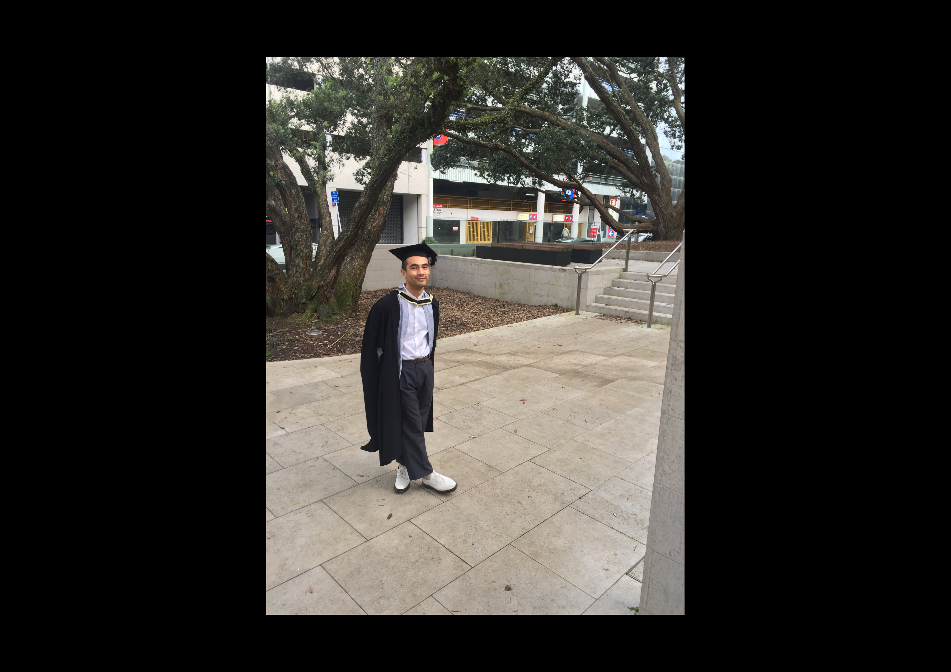 Micheal wears a graduation cap and gown and is photographed in a university courtyard.