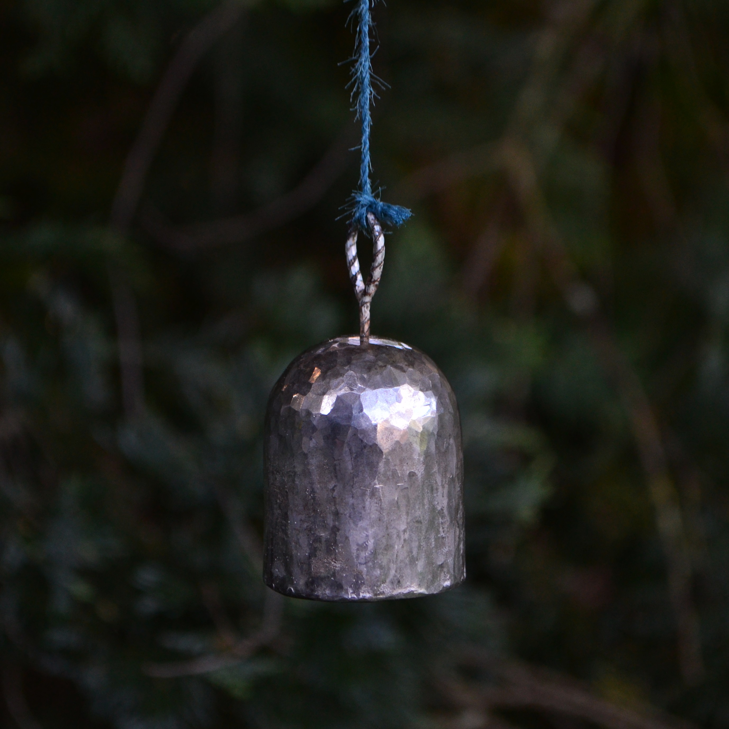 A hand-beaten silver bell hanging on an indigo cord.