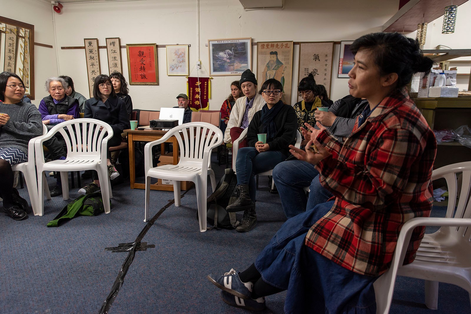 Kerry Ann Lee speaks in the foreground as other Asian people watch on, inside Chinese community rooms.