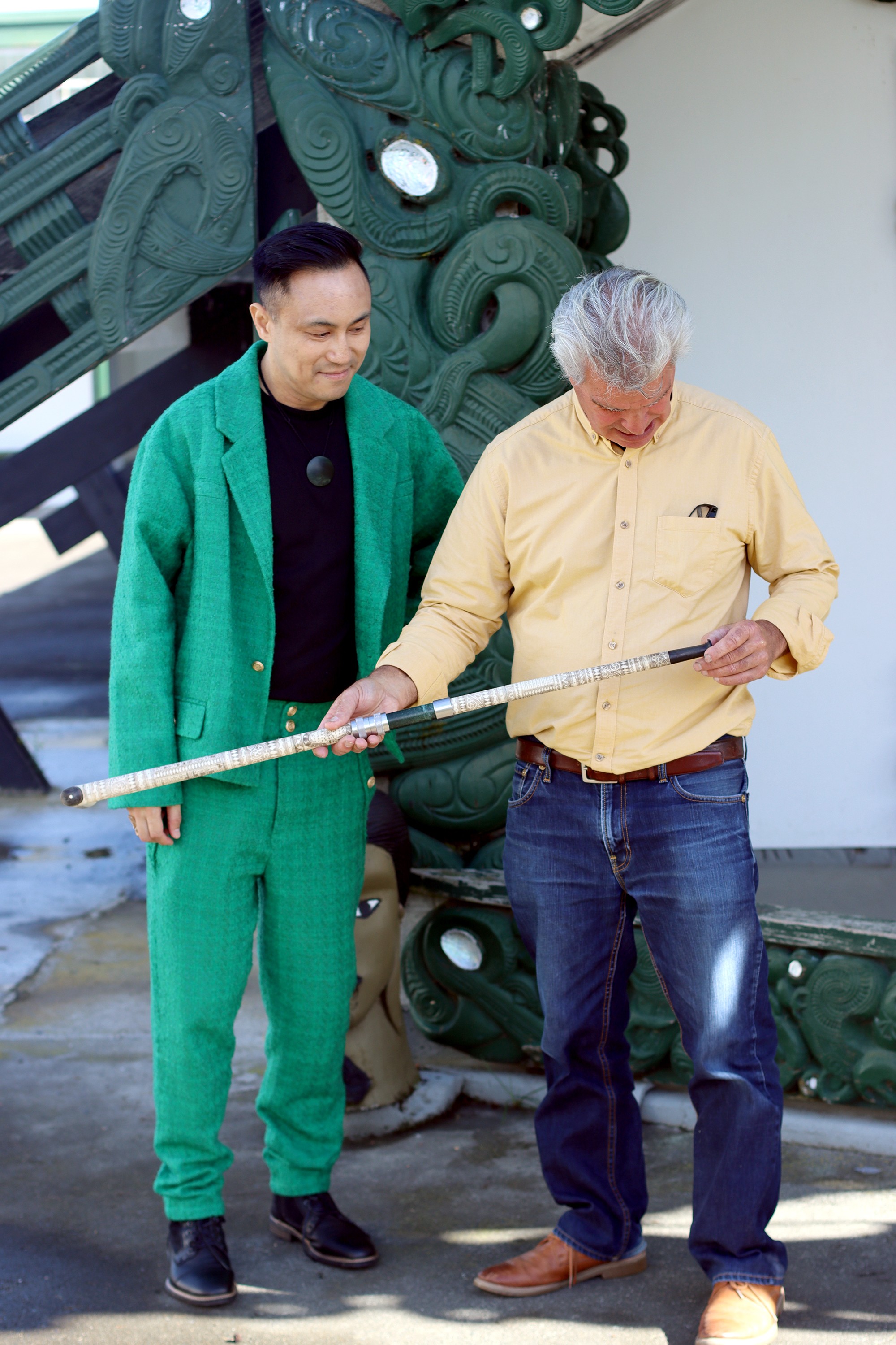 Chris Tse wears a green suit and stands next to carver Jacob Scott who is holding a tokotoko that they are both admiring.