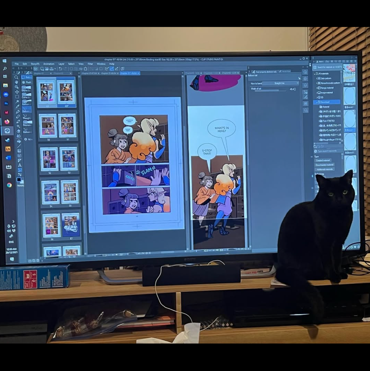 A photograph of a computer with comic pages laid out in a design software. There is a black cat sitting to the right of the computer. 