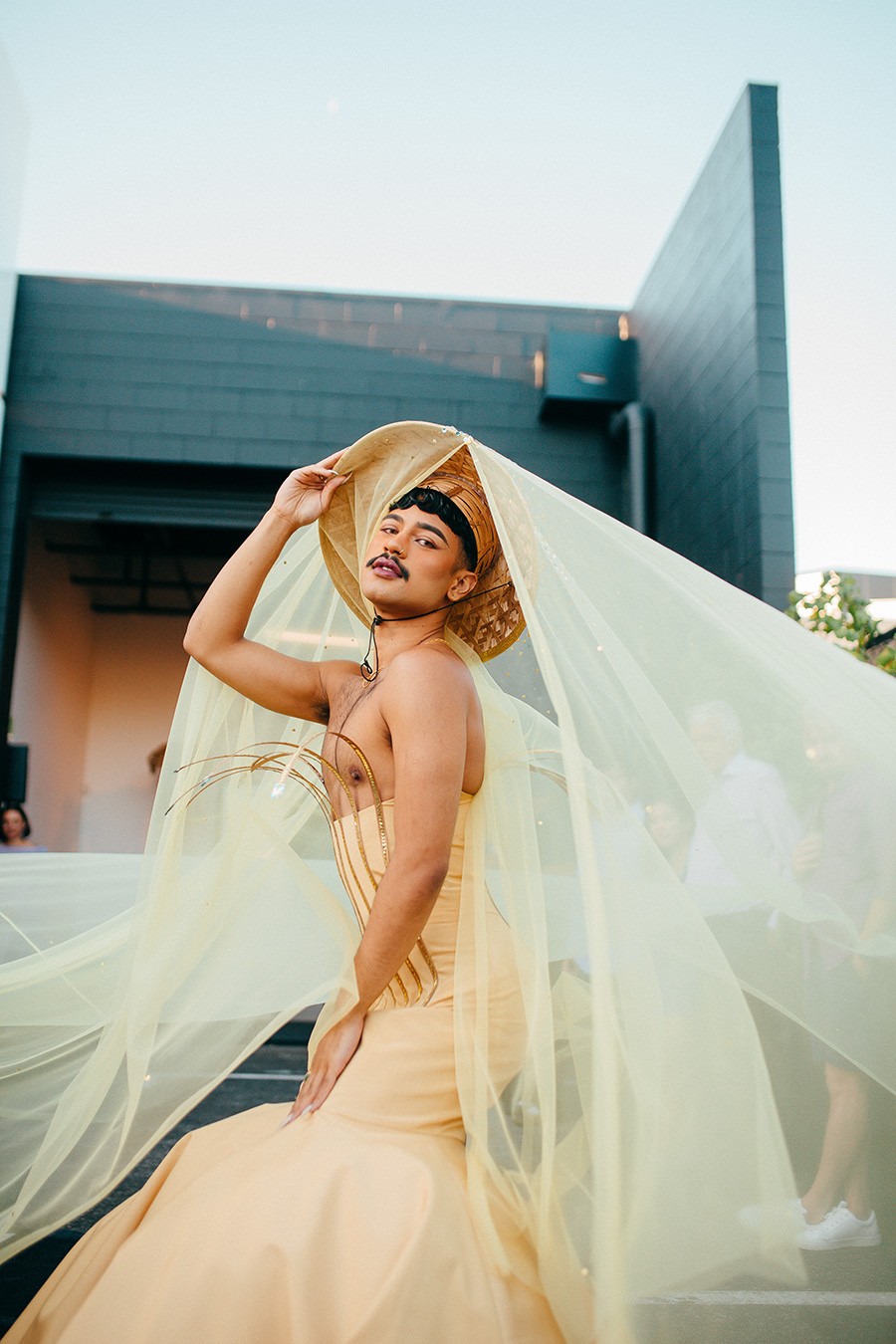 Person wearing a golden dress that shows off their chest and a wide brimmed hat with flowing golden fabric