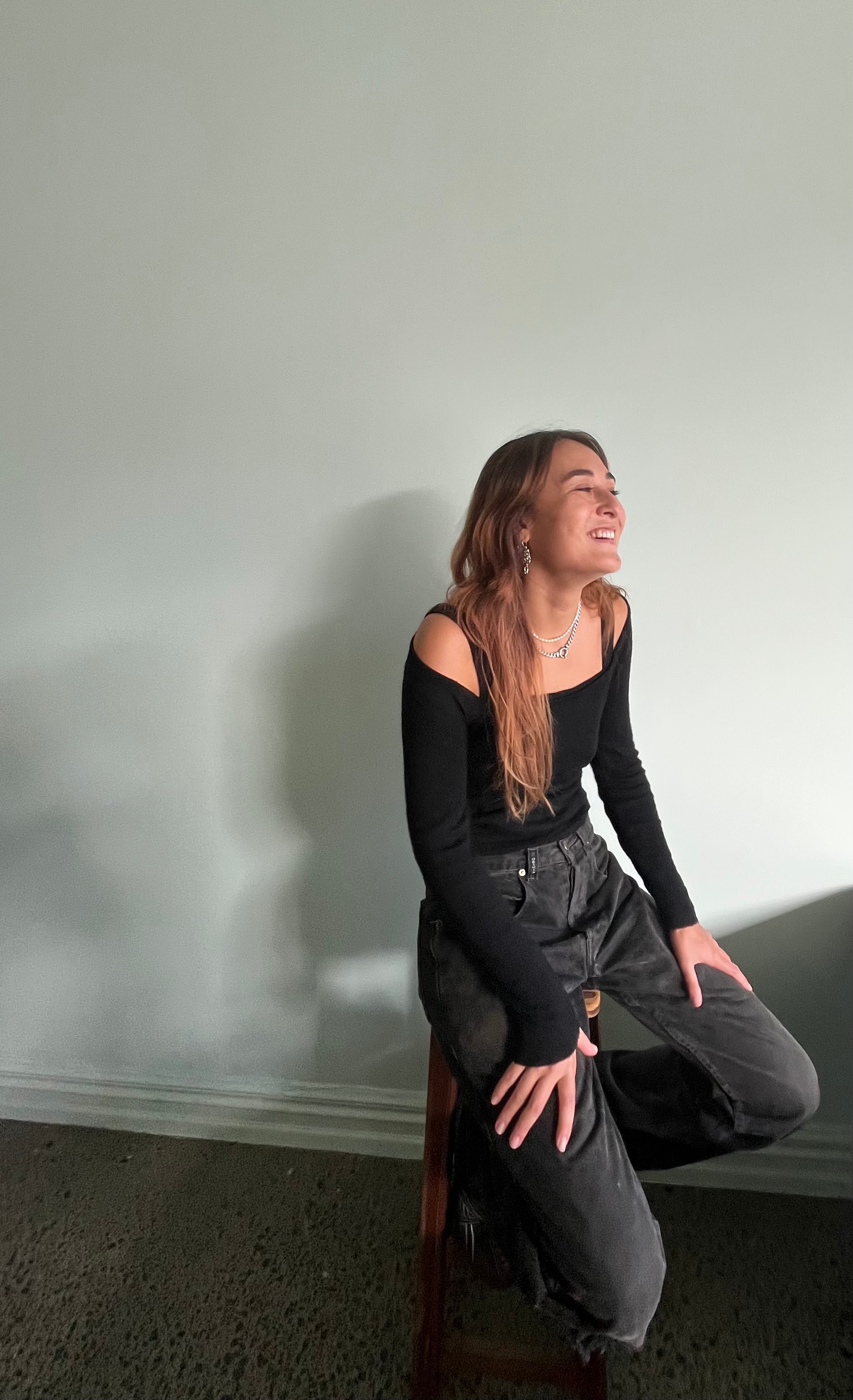 A woman with long hair sits on a stool and is smiling at someone outside of the photo