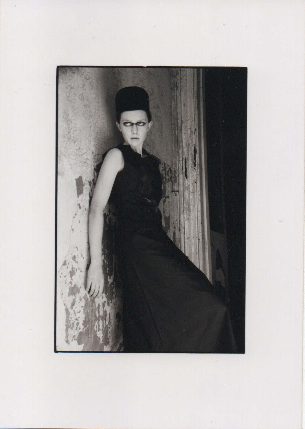 A black and white photo of a woman in a draped floor-length outfit leaning against a stone wall