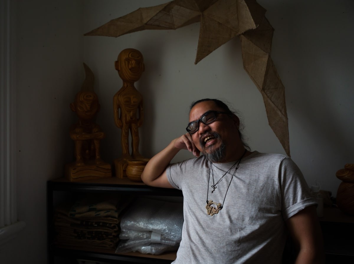 A Filipino smiling wearing a grey shirt and a copper pendant in a dimly-lit studio surrounded by various brown sculptures