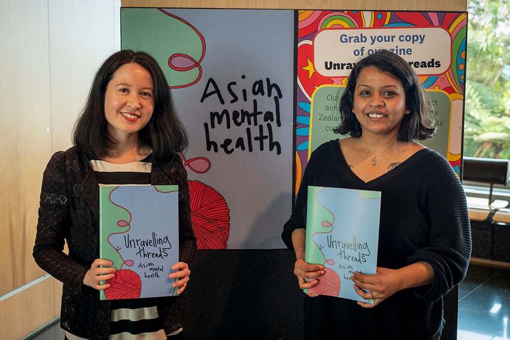 Two women smiling next to a blue poster that says 'Asian Mental Health'.