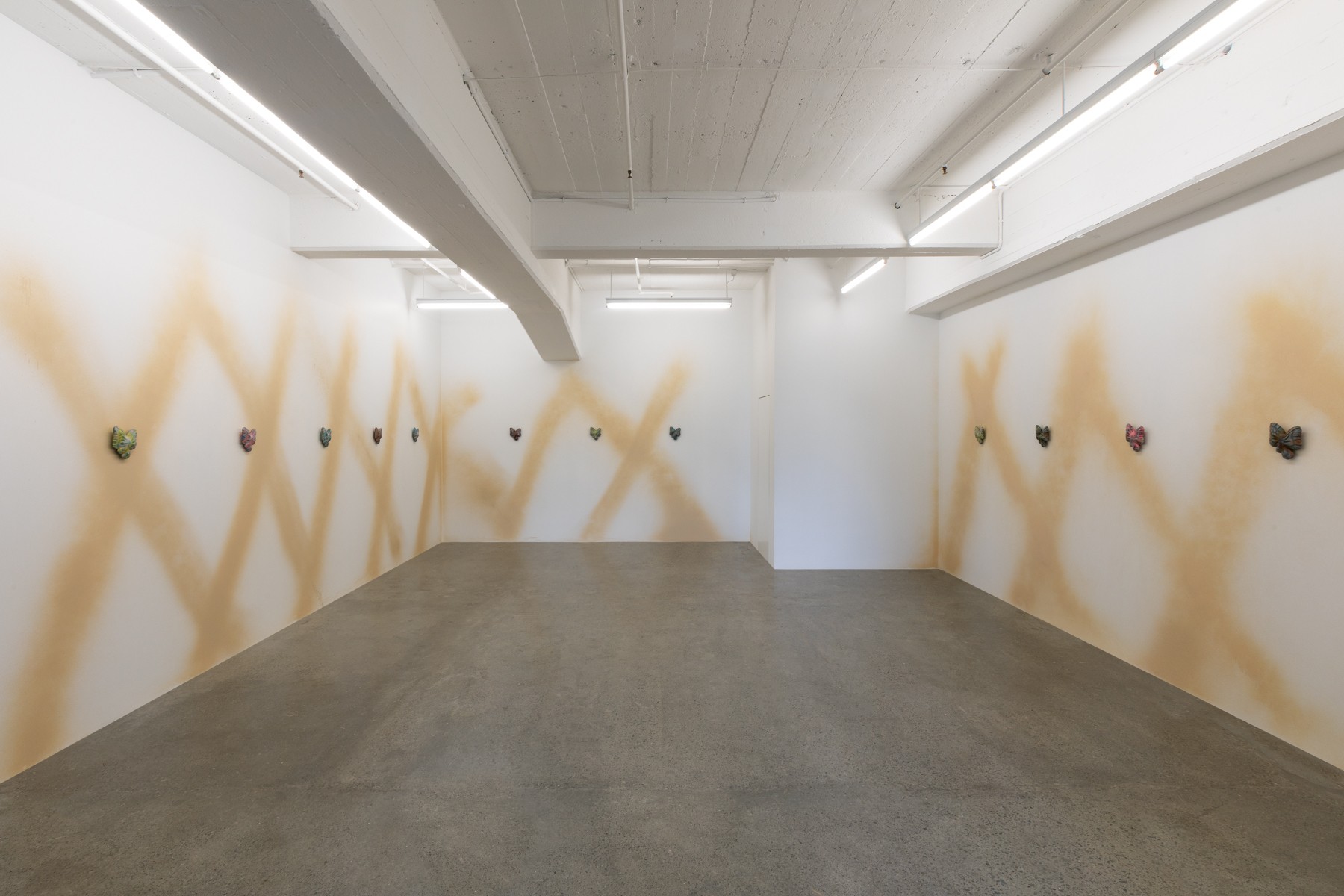 A white-walled gallery with light brown X's spraypainted across the full wall, almost resembling a gate, with cast butterflies mounted in the centre of the wall at regular intervals