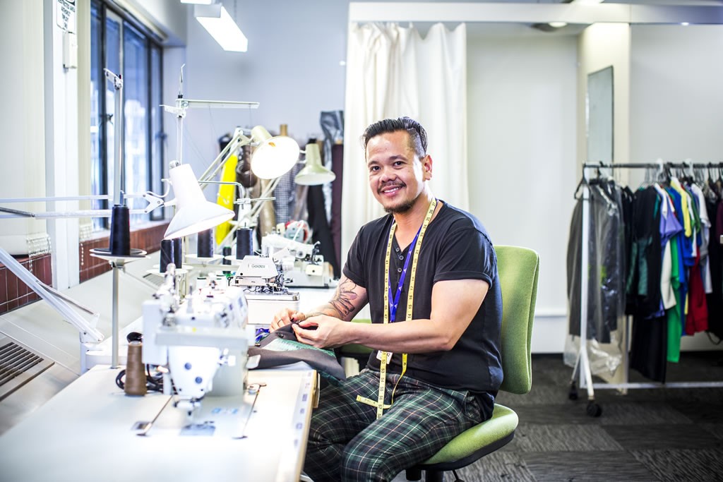 A similing FIlipino wearing a black top and checkered pants with a tape measure around their neck sitting down on a sewing machine table working on grey fabric