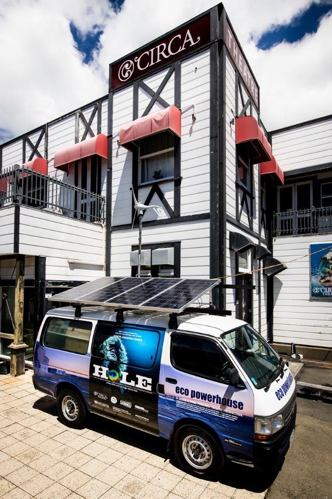 A white van is parked outside of a theatre with artwork advertising a play on its side. Solar panels are installed on top of the van.