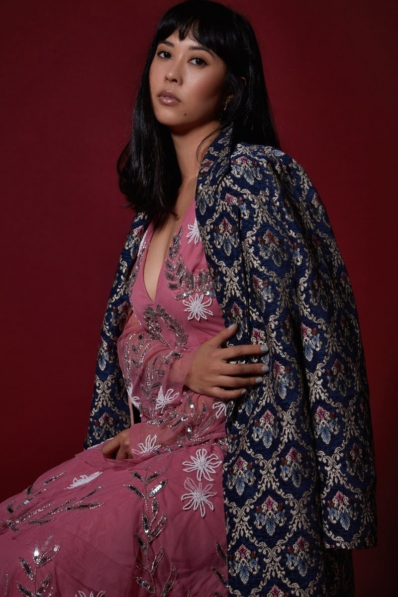 A woman with shoulder-length black hair and a pink embroidered dress with a jacquard jacket resting on her shoulders sits on a stool gazing at the camera