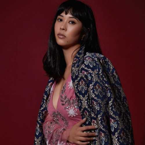 A woman with shoulder-length black hair and a pink embroidered dress with a jacquard jacket resting on her shoulders sits on a stool gazing at the camera