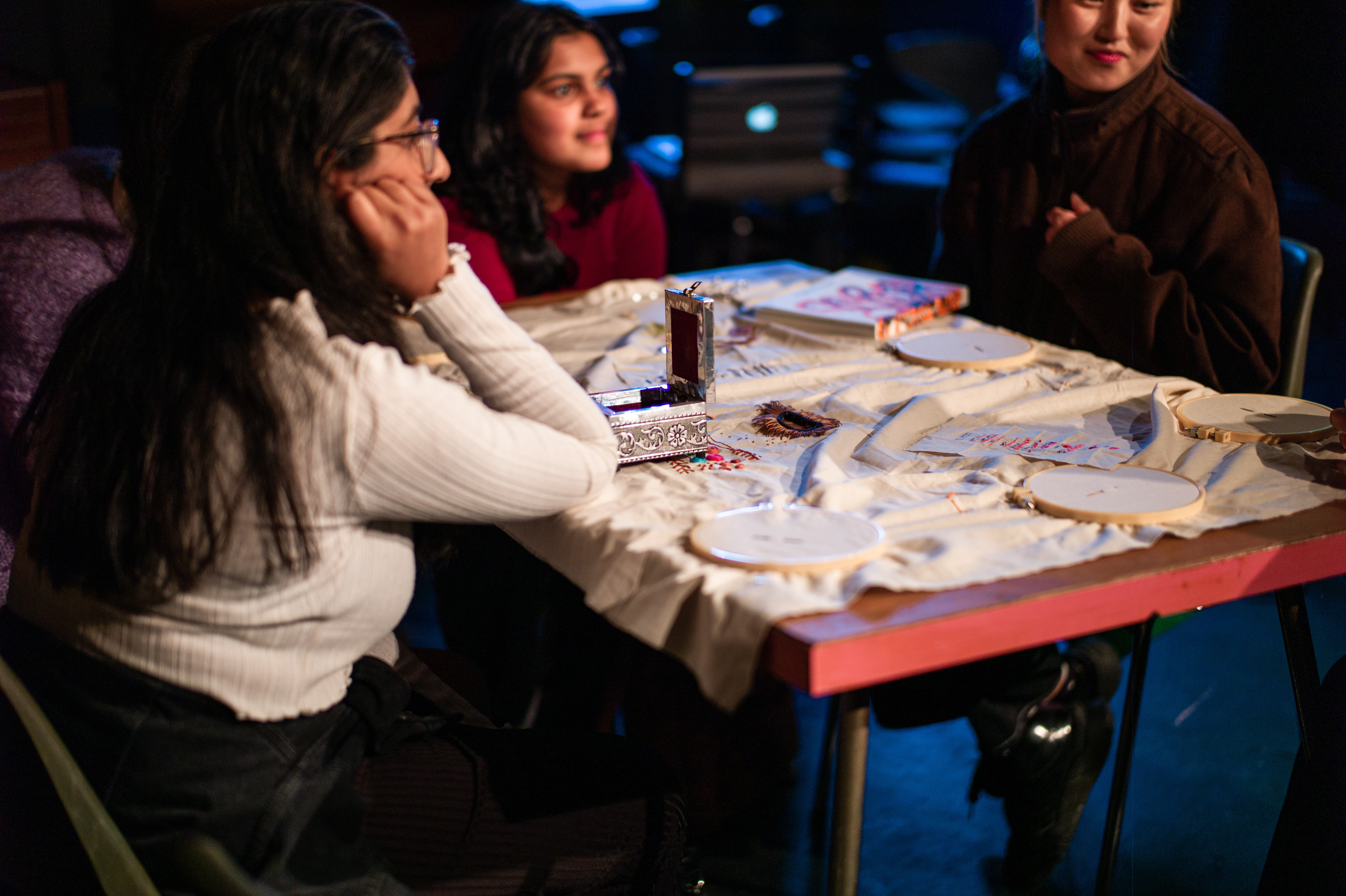 Three women sitting around a table with a piece of fabric with embroidery materials covering the table. 