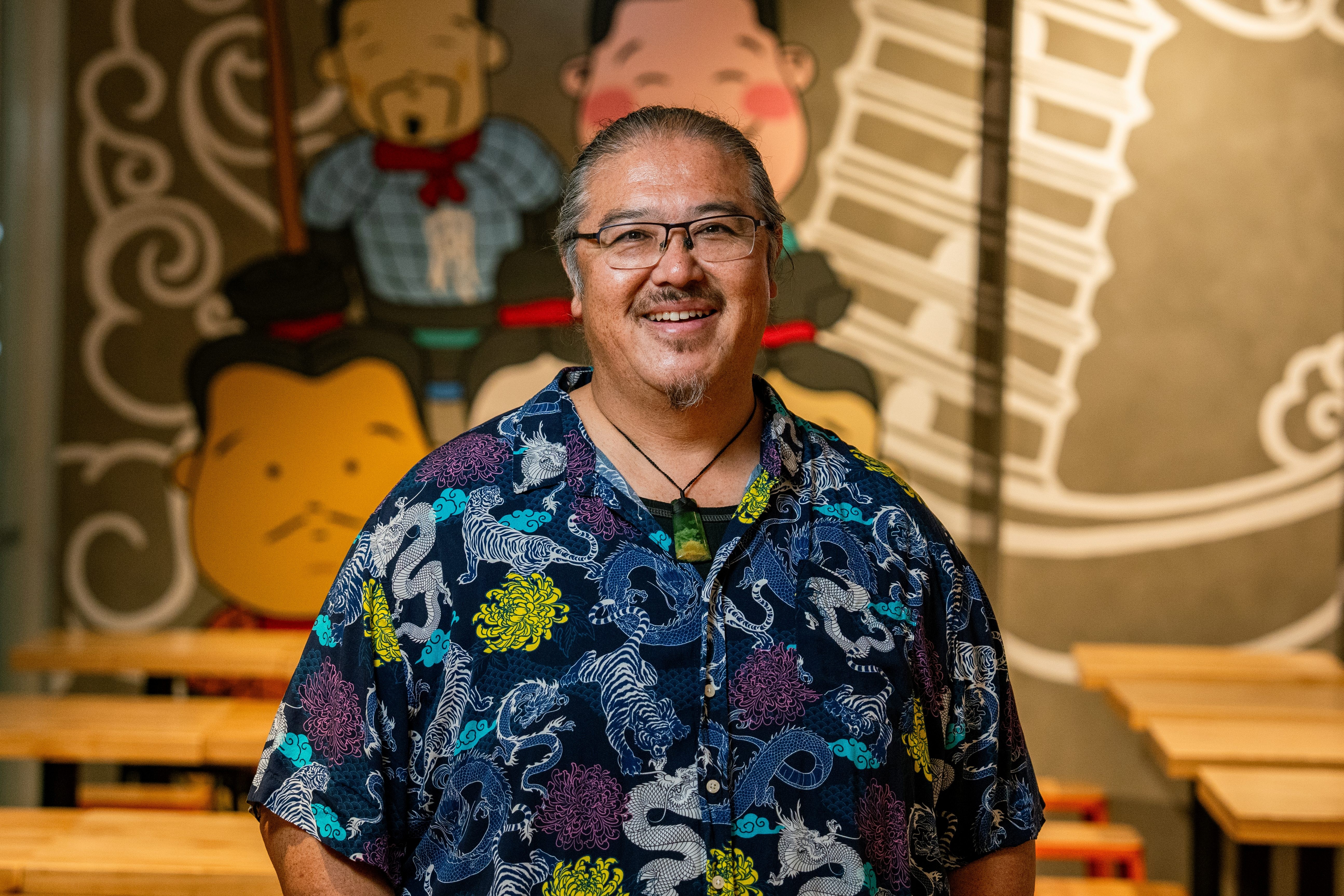 A smiling man with multi-coloured shirt on standing in a restaurant.