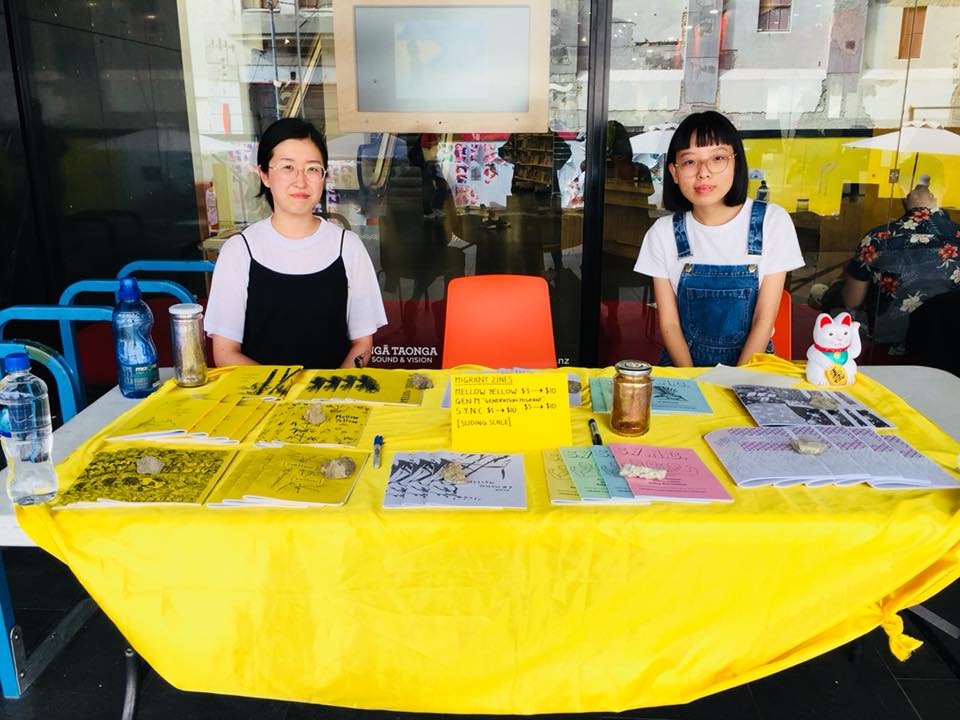 Two people smiling and sitting behind a table displaying various zines on top of a yellow tablecloth. 
